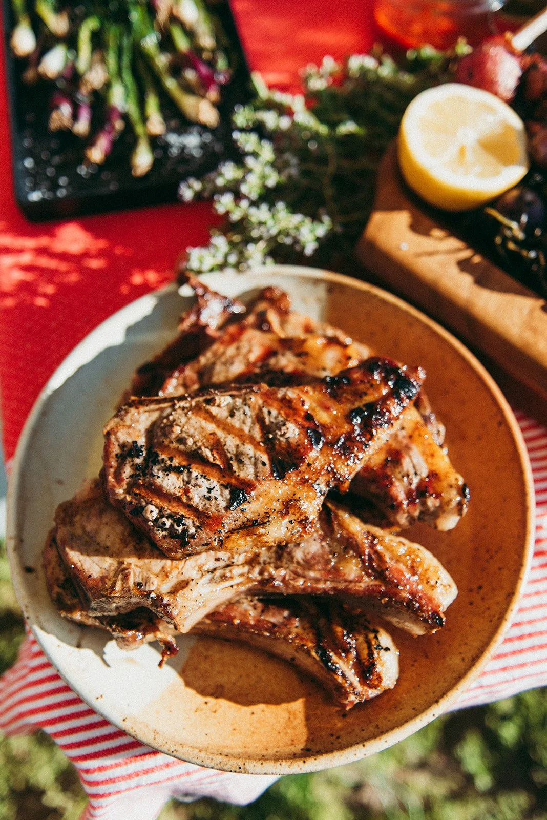 Grilled pork chops on a beige plate with a balsamic glaze, with a side of lemon and greens, on a red-striped cloth, outdoors.