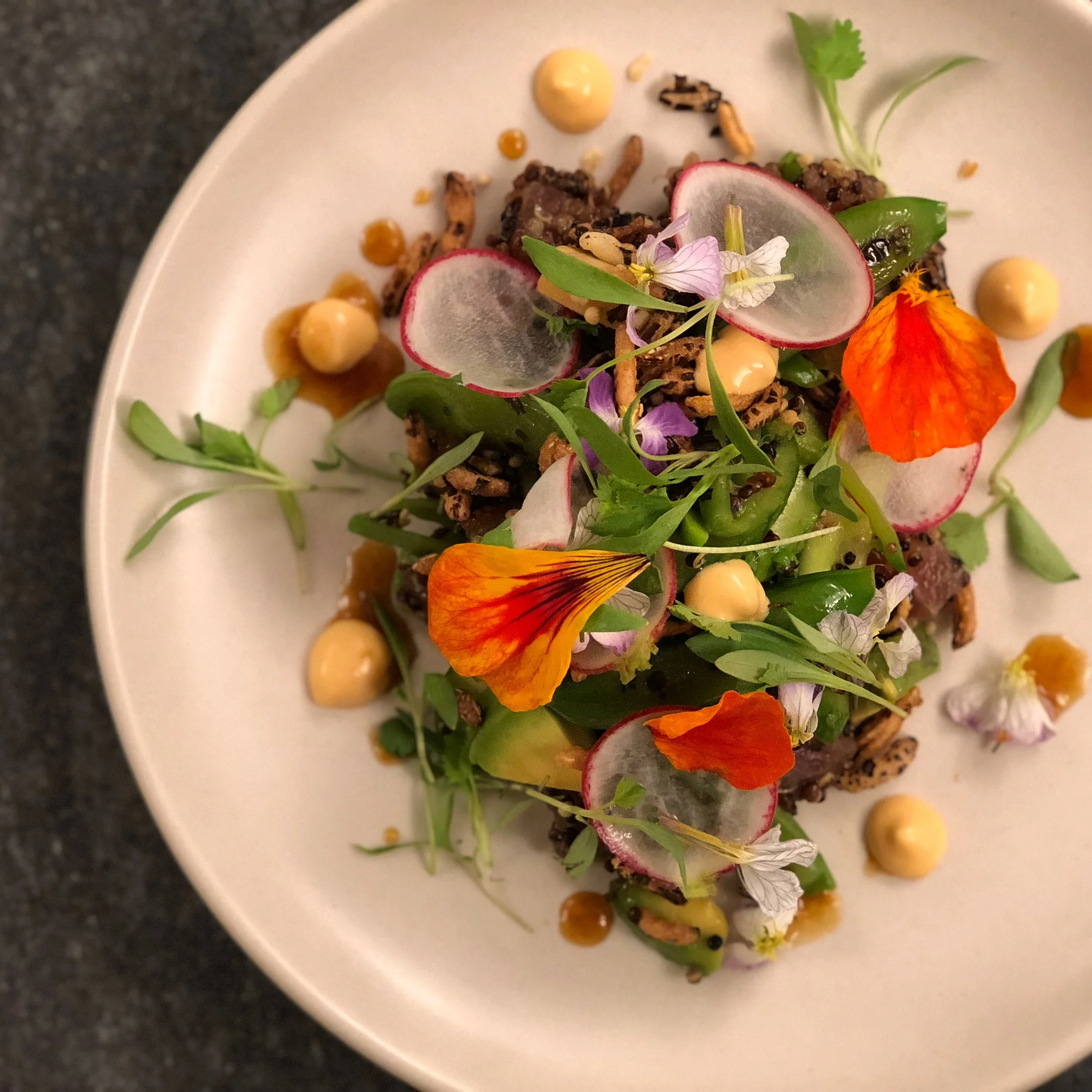 A colorful plated dish with radish slices, edible flowers, microgreens, and a creamy sauce or dressing, garnished decoratively.