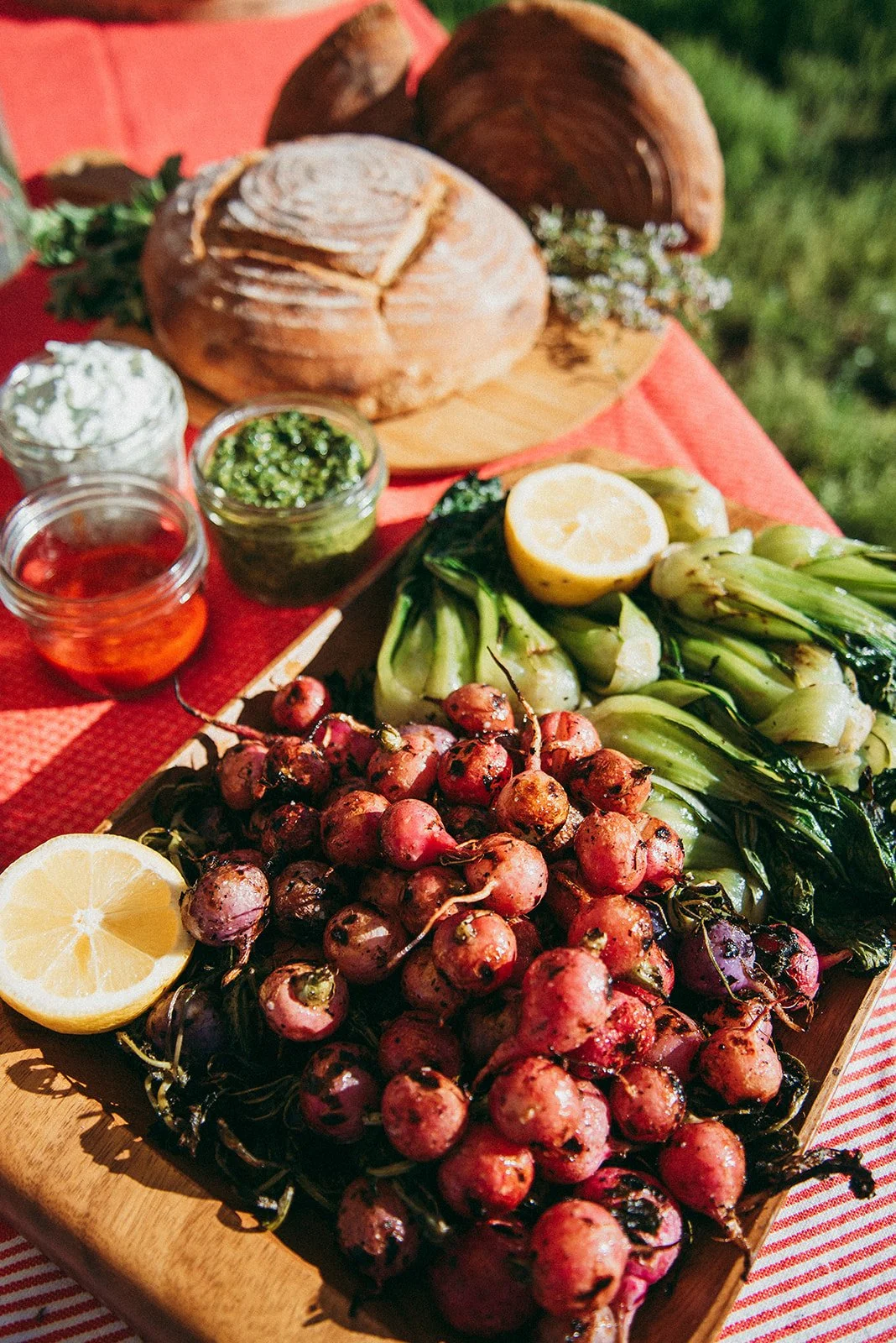 A wooden tray with fresh radishes, grilled bok choy, and lemon halves, set on a red-striped tablecloth. In the background, there is a loaf of crusty bread, small jars of herbs, jam, and cream, and a sprig of herbs, all outdoors on a sunny day.