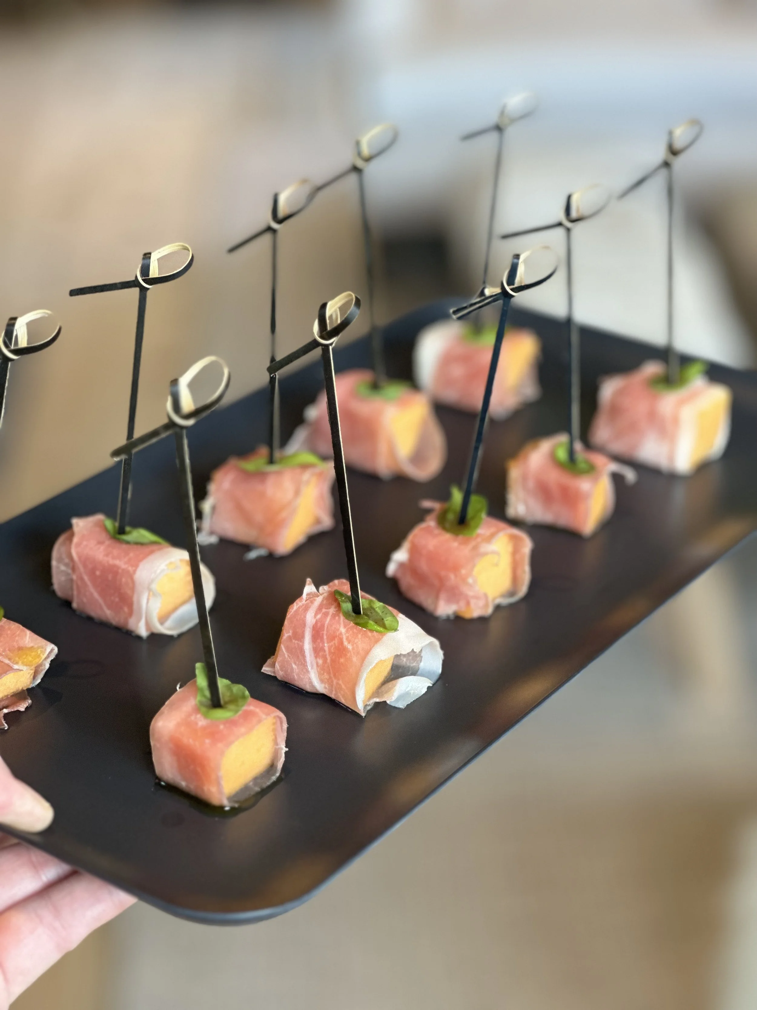 Appetizer bites with prosciutto, cheese, and greens secured with black skewers on a black serving tray.