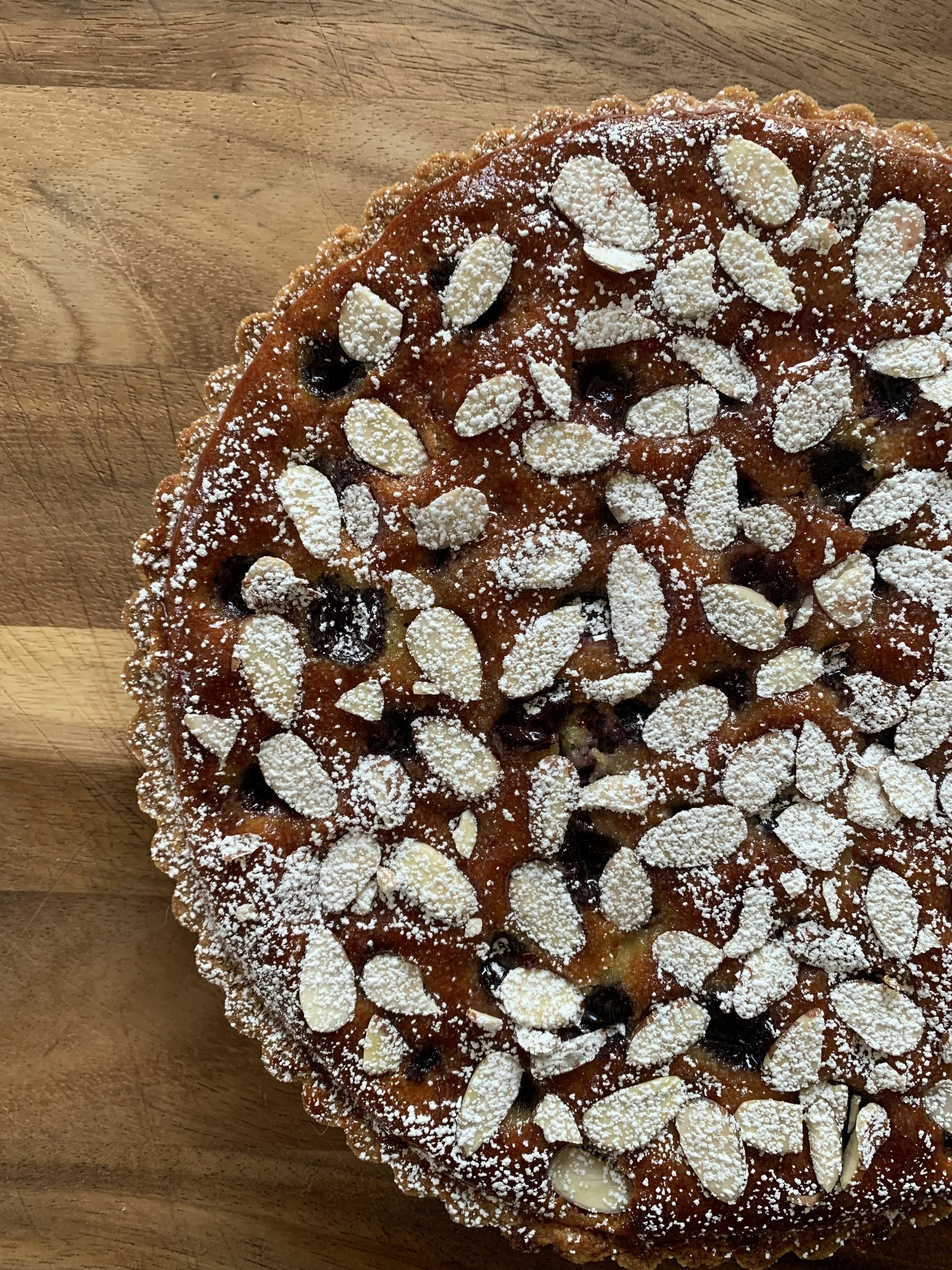 A round tart topped with sliced almonds, powdered sugar, and blueberries, sitting on a wooden surface.