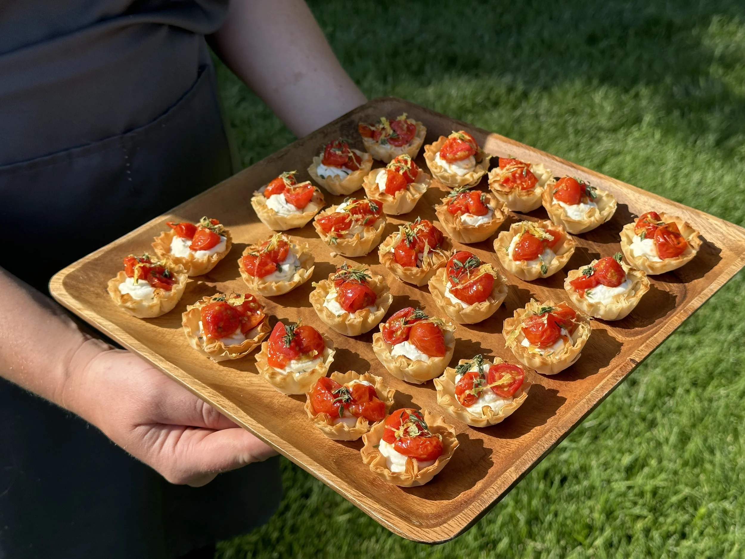 A person holding a wooden tray with bite-sized appetizer tartlets topped with cherry tomatoes, basil, and cream cheese, outdoors on a grassy area.