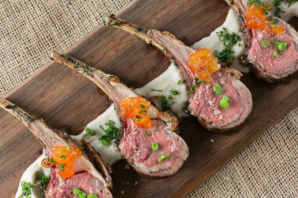 Four cooked rack of lamb chops served with a white sauce, garnished with chopped green onions and orange-colored sauce or garnish on top of each.