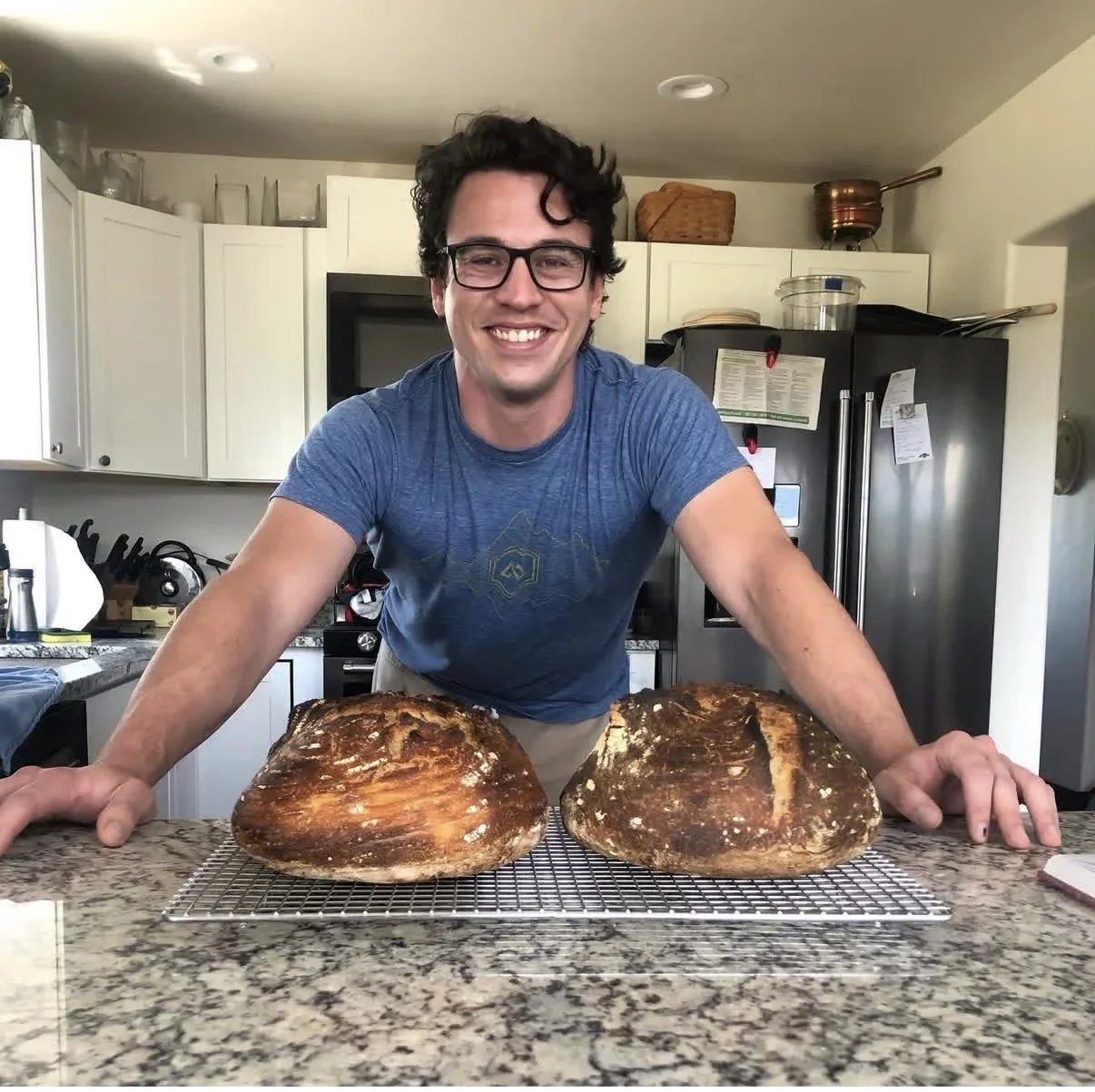 A man smiling, wearing glasses and a blue t-shirt, leaning on a kitchen counter with two large, baked bread loaves cooling on a wire rack in front of him.