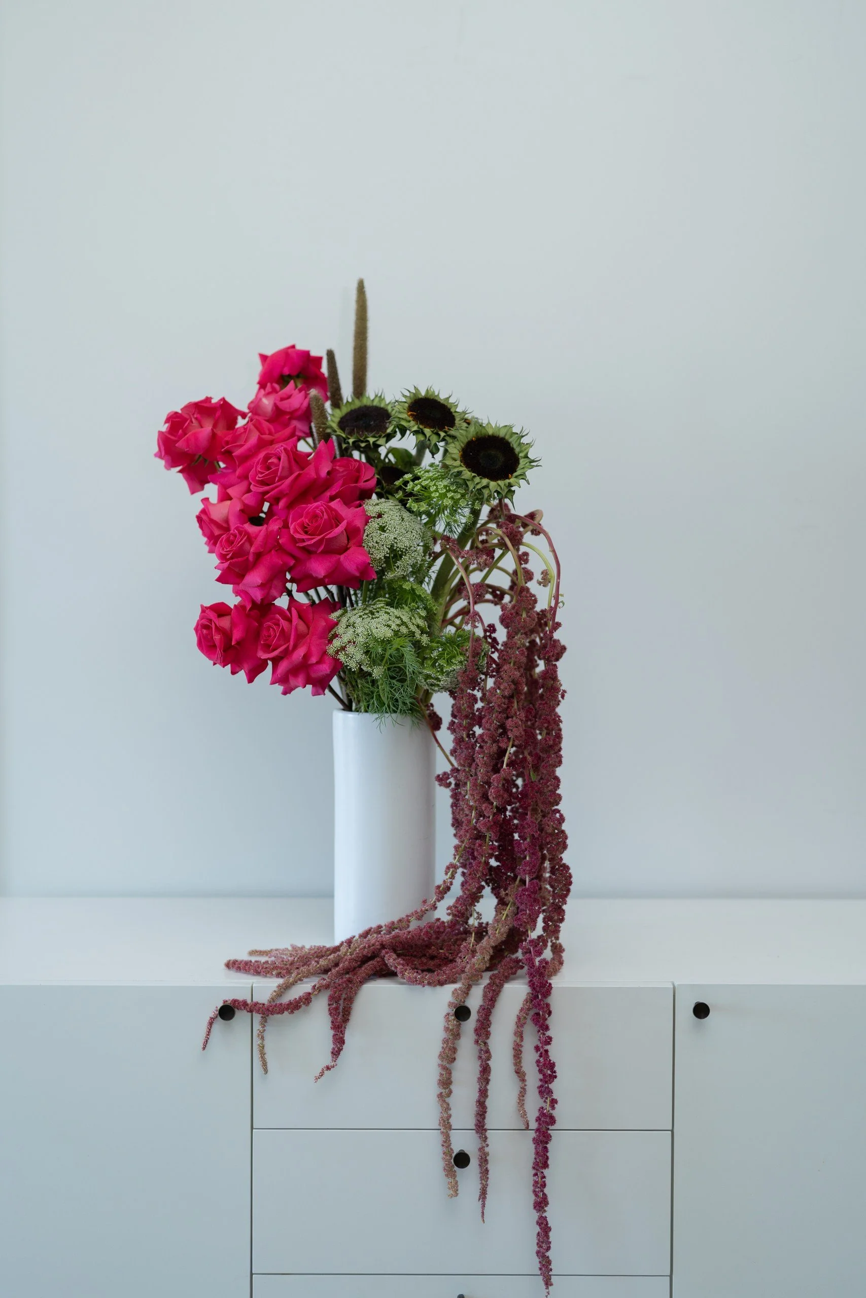 Magenta roses, green flowers and trailing burgundy amaranthus in a tall white vase.