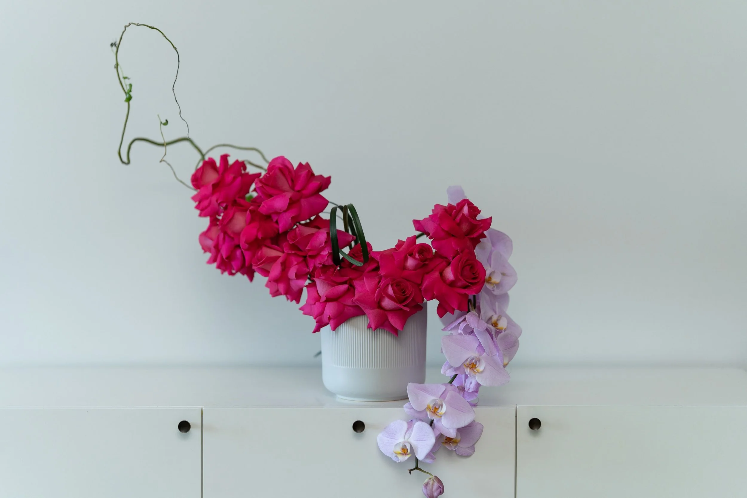 Magenta roses, pale orchids and curling branches in a white ceramic vase.