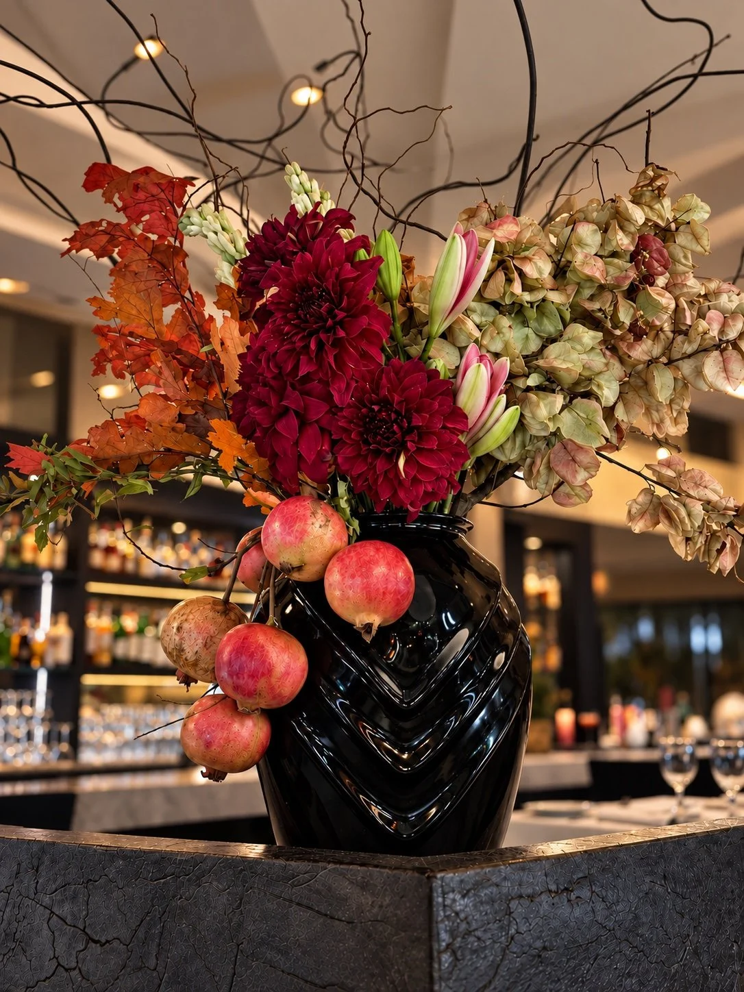 Sculptural flowers, branches and pomegranates in a black vase within a hospitality interior.