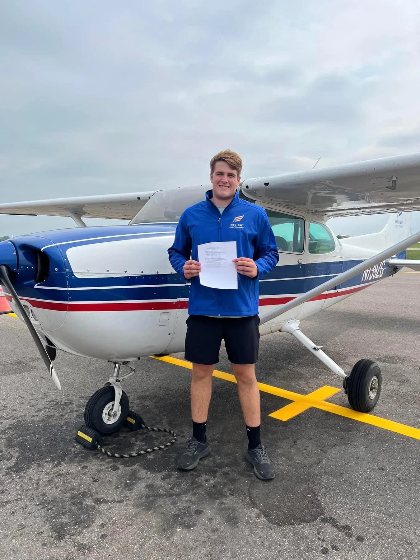 Young man in a blue flight suit standing in front of a small airplane on a tarmac, holding a certificate, with a grip and rope on the ground nearby.