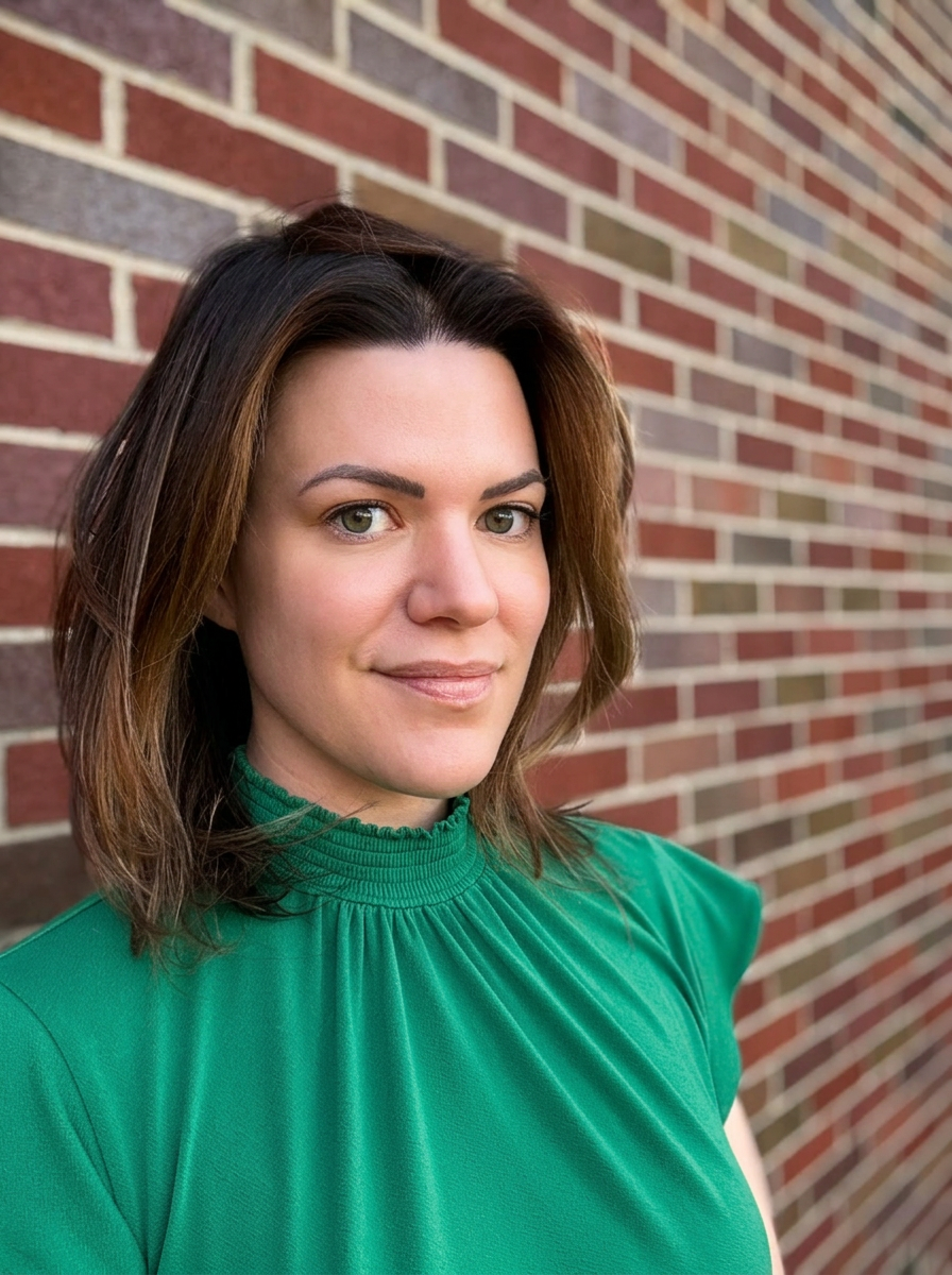 A woman with shoulder-length brown hair, green eyes, and light skin wearing a green blouse standing next to a brick wall.