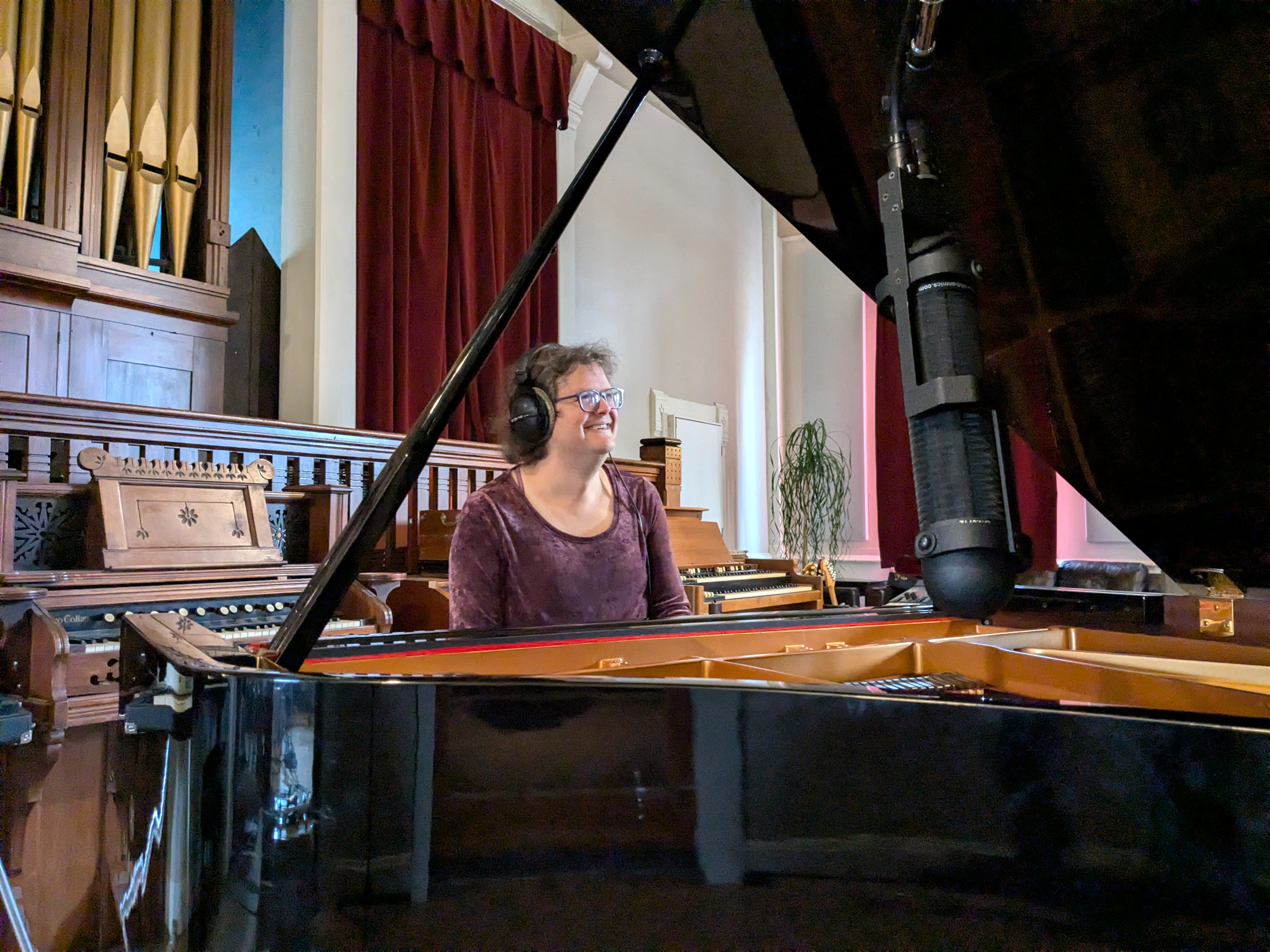 A person with glasses and wearing headphones sitting at a grand piano, in a room with red curtains, a wooden organ with pipes, and a plant in a white pot.