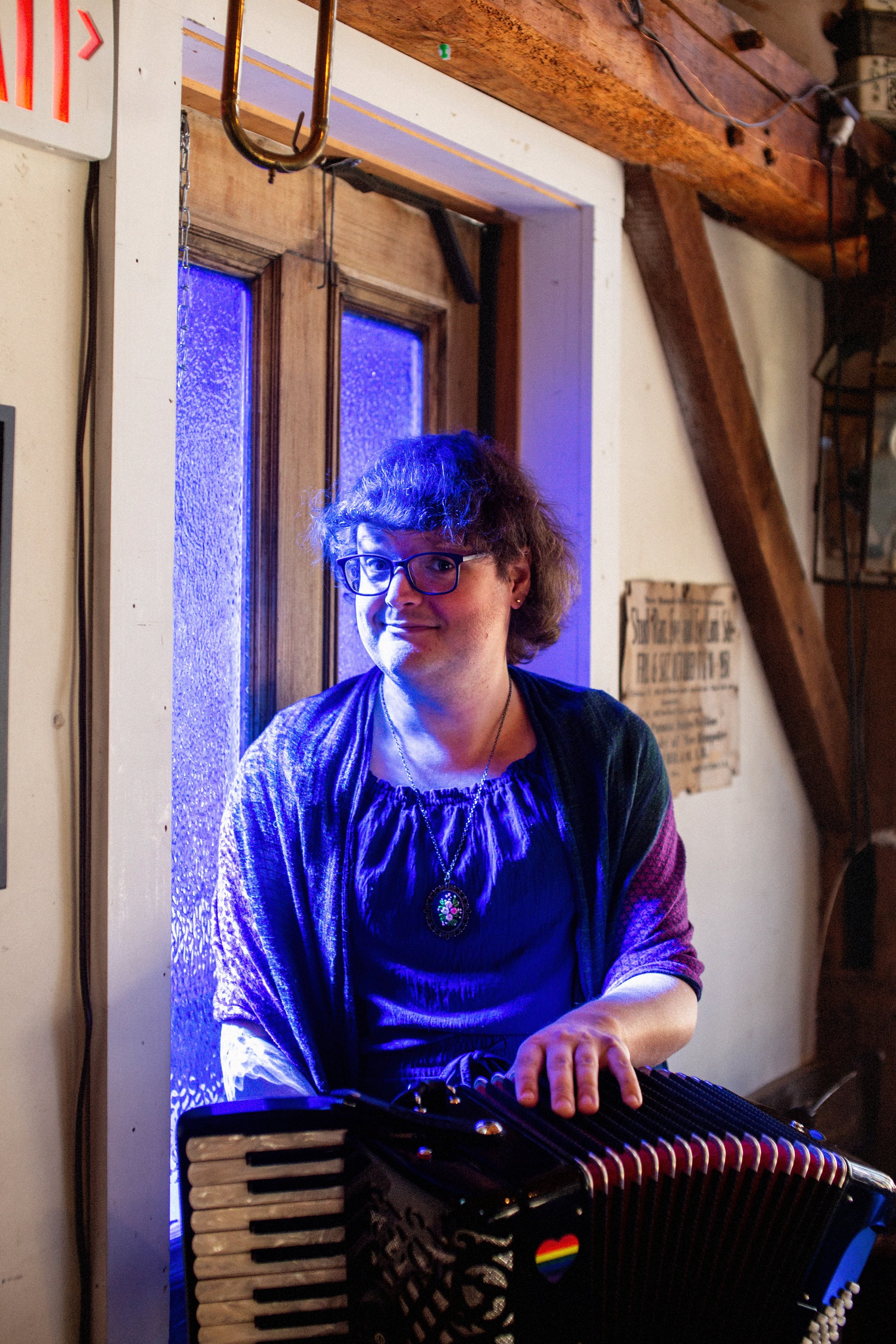A woman with glasses and curly hair playing an accordion in a cozy, rustic room with wooden beams and a window with blue-tinted glass.