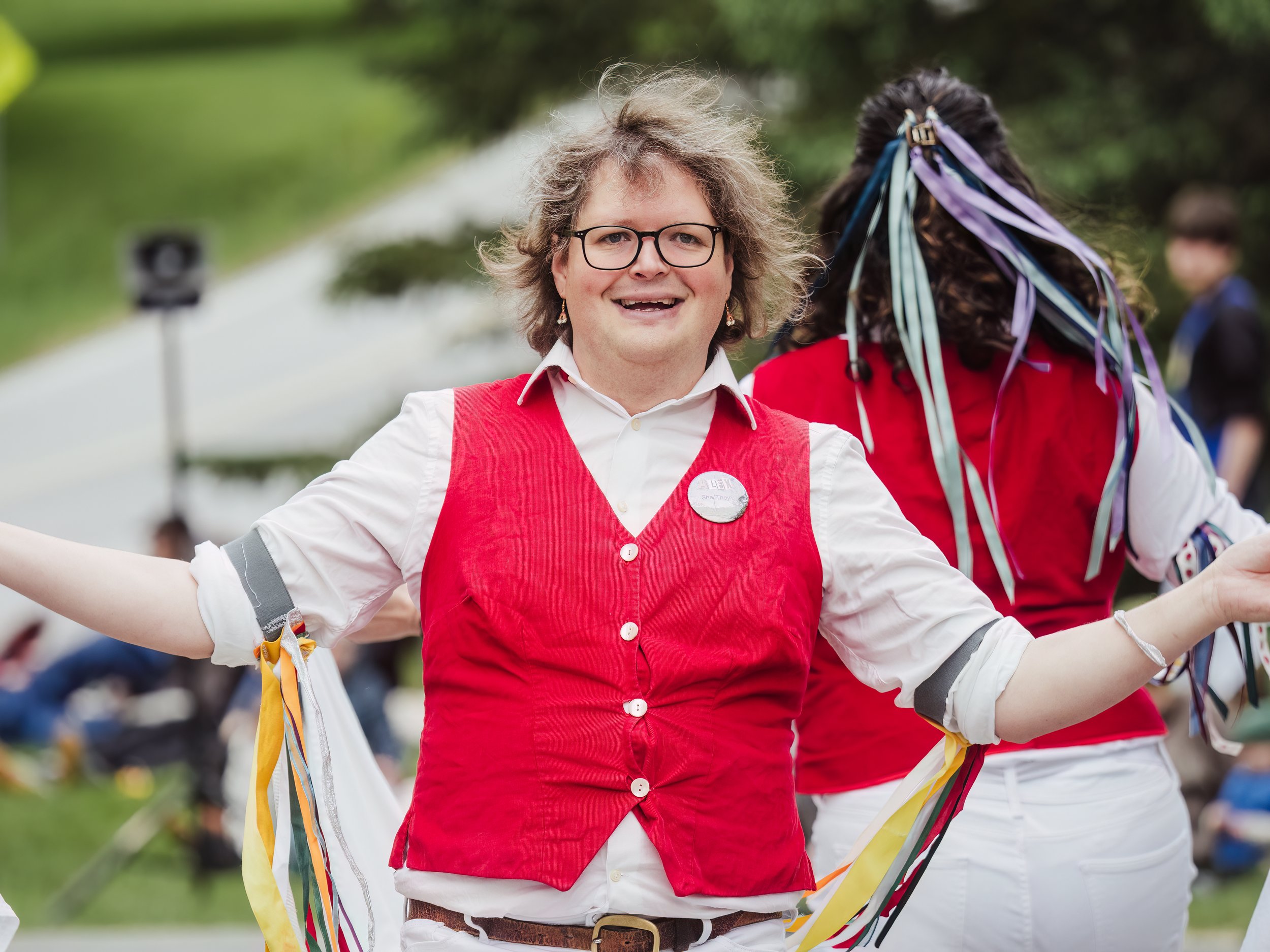 Person with curly hair, glasses, and a red vest smiling at an outdoor event, with colorful ribbons in the background.