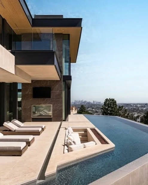 Modern house with a rooftop infinity pool overlooking a city skyline, with lounge chairs and a seating area.