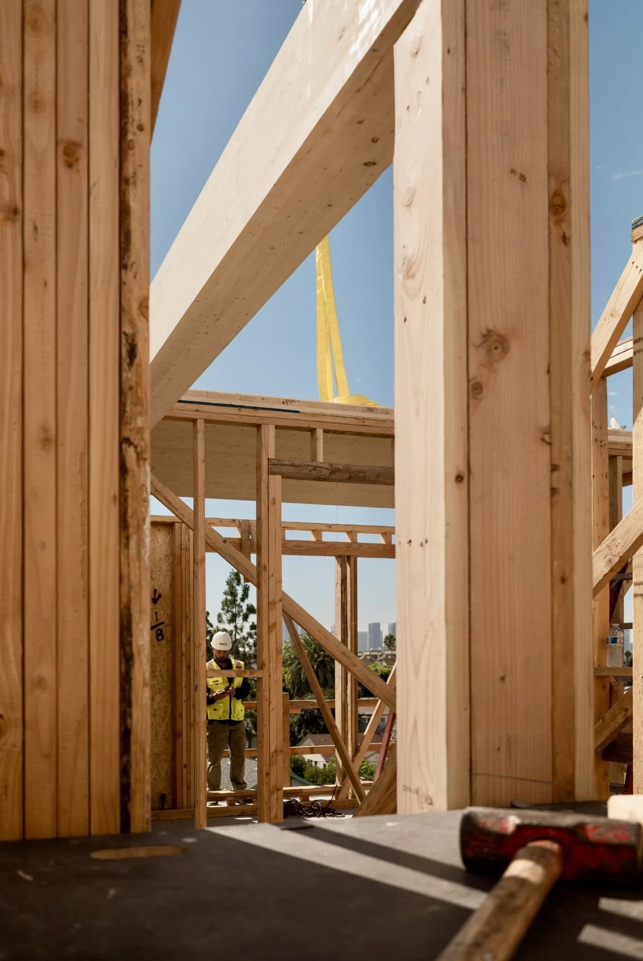 Construction site with wooden framing, a worker wearing a white helmet and yellow vest is seen in the background, and construction tools are on a surface in the foreground.