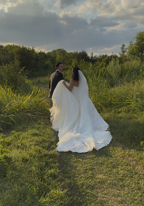 Couple dressed in wedding attire holding hands in a grassy outdoor setting with trees and a cloudy sky in the background. Love Letty Content, Luxury Wedding Content Creation in Dallas Texas.