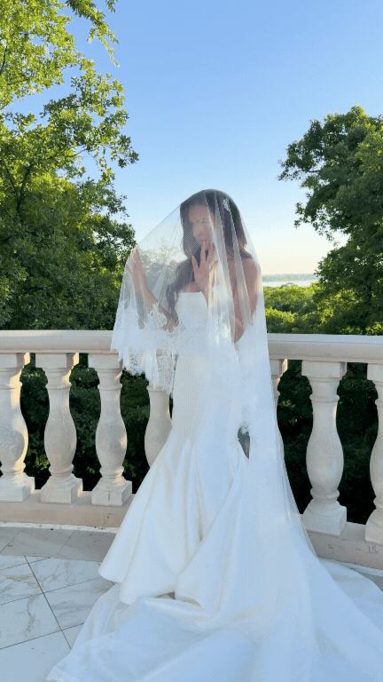 A woman in a white wedding dress standing on a balcony with a lace veil over her face. She is outside, surrounded by green trees, with a clear blue sky in the background. Love Letty Content, Luxury Wedding Content Creation in Dallas Texas.