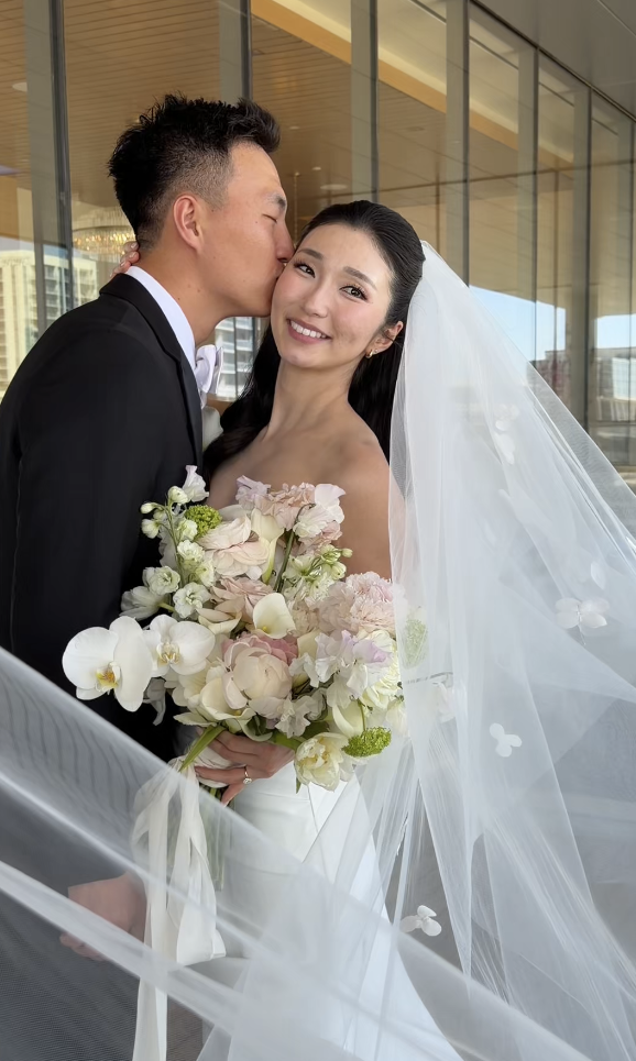 A bride and groom sharing a moment indoors, with the groom kissing the bride on the cheek. The bride is holding a bouquet of white and light pink flowers, and she is wearing a white wedding dress with a veil. Large windows in the background reveal a cityscape.