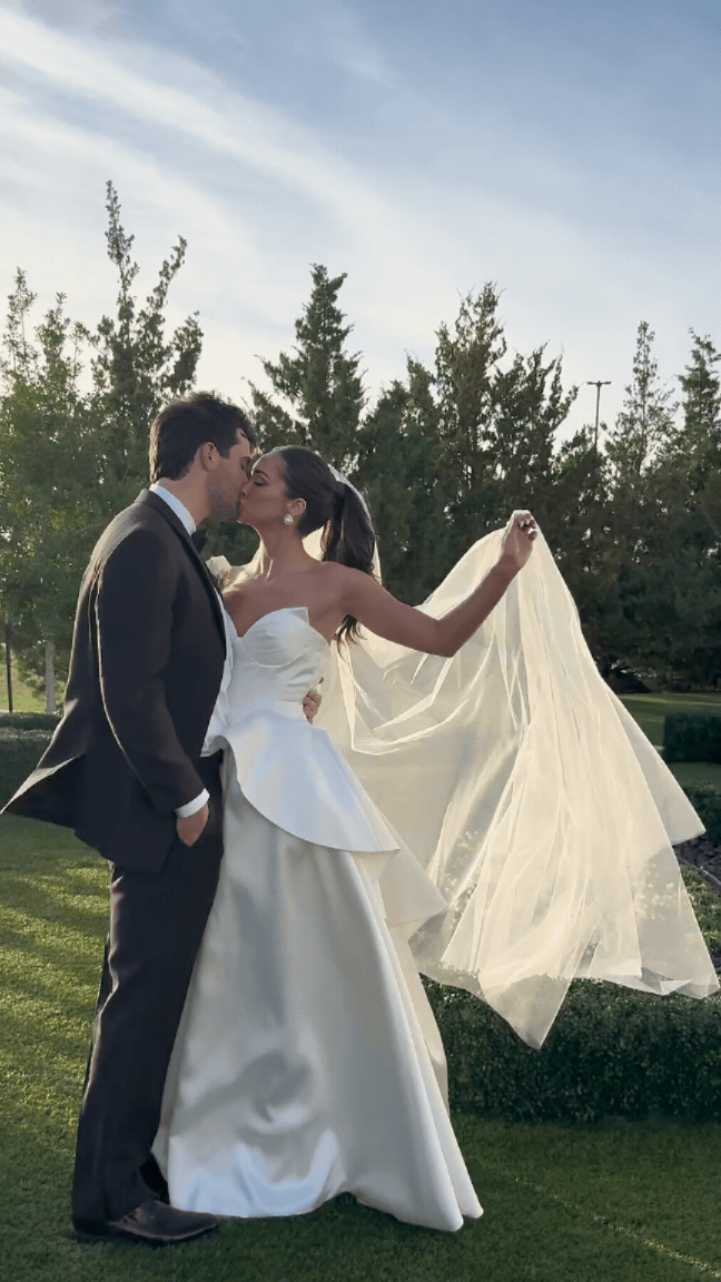 A bride and groom sharing a kiss outdoors, with the bride holding up her wedding veil, against a backdrop of trees and a partly cloudy sky. Love Letty Content, Luxury Wedding Content Creation in Dallas Texas.