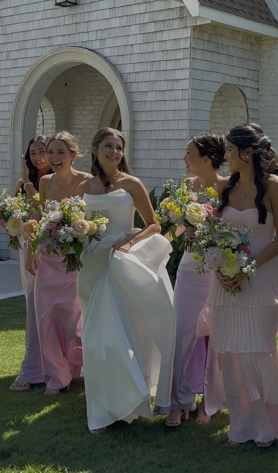 A bride and five women in pastel dresses holding bouquets of flowers walking outside near a house with white brick walls. Love Letty Content, Luxury Wedding Content Creation in Dallas Texas.