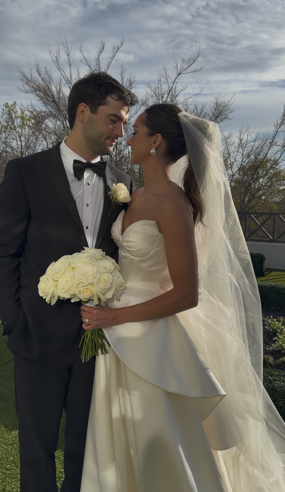 Bride and groom standing close, looking at each other, outdoors on a wedding day. The bride wears a strapless white wedding gown and veil, holding a bouquet of white roses. The groom wears a black tuxedo with a bow tie and boutonniere. Trees and a cloudy sky are in the background.