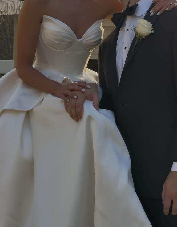 A bride in a white strapless wedding gown sitting with her hands resting on the groom's hand, who is wearing a dark tuxedo with a white shirt, black bow tie, and white flower boutonniere, at a wedding event.