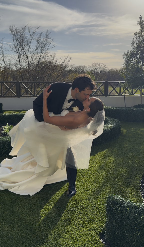 A newlywed couple sharing a kiss outdoors during their wedding celebration, with the groom dipping the bride against a backdrop of trees and a wooden fence on a sunny day.