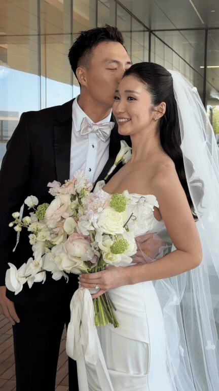 A bride and groom standing close together, with the groom kissing the bride on the temple. The bride is smiling and holding a large bouquet of flowers. They are outdoors, near a building with glass walls. Love Letty Content, Luxury Wedding Content Cr