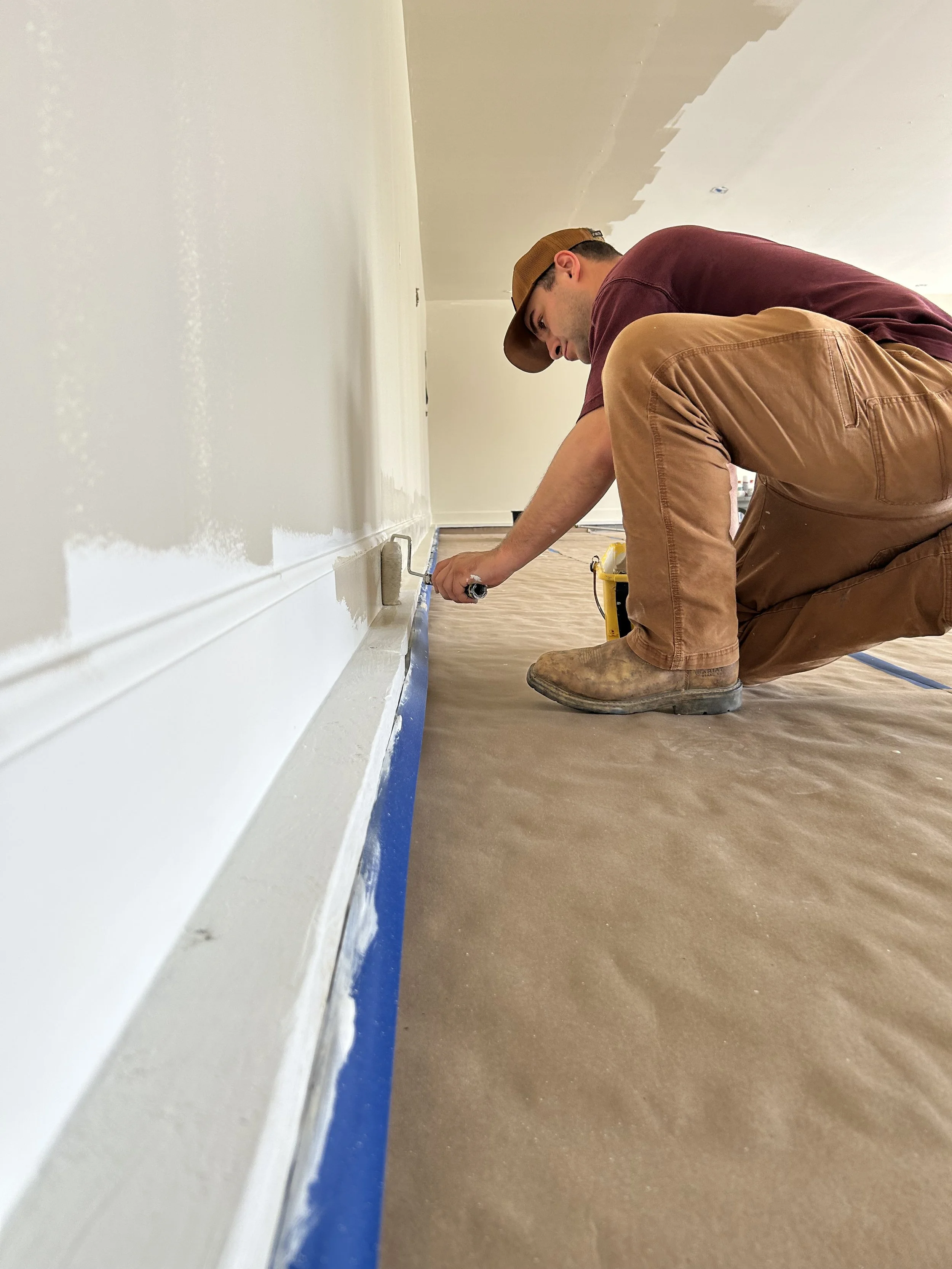 A man kneeling on a floor painting a baseboard with a paint roller.