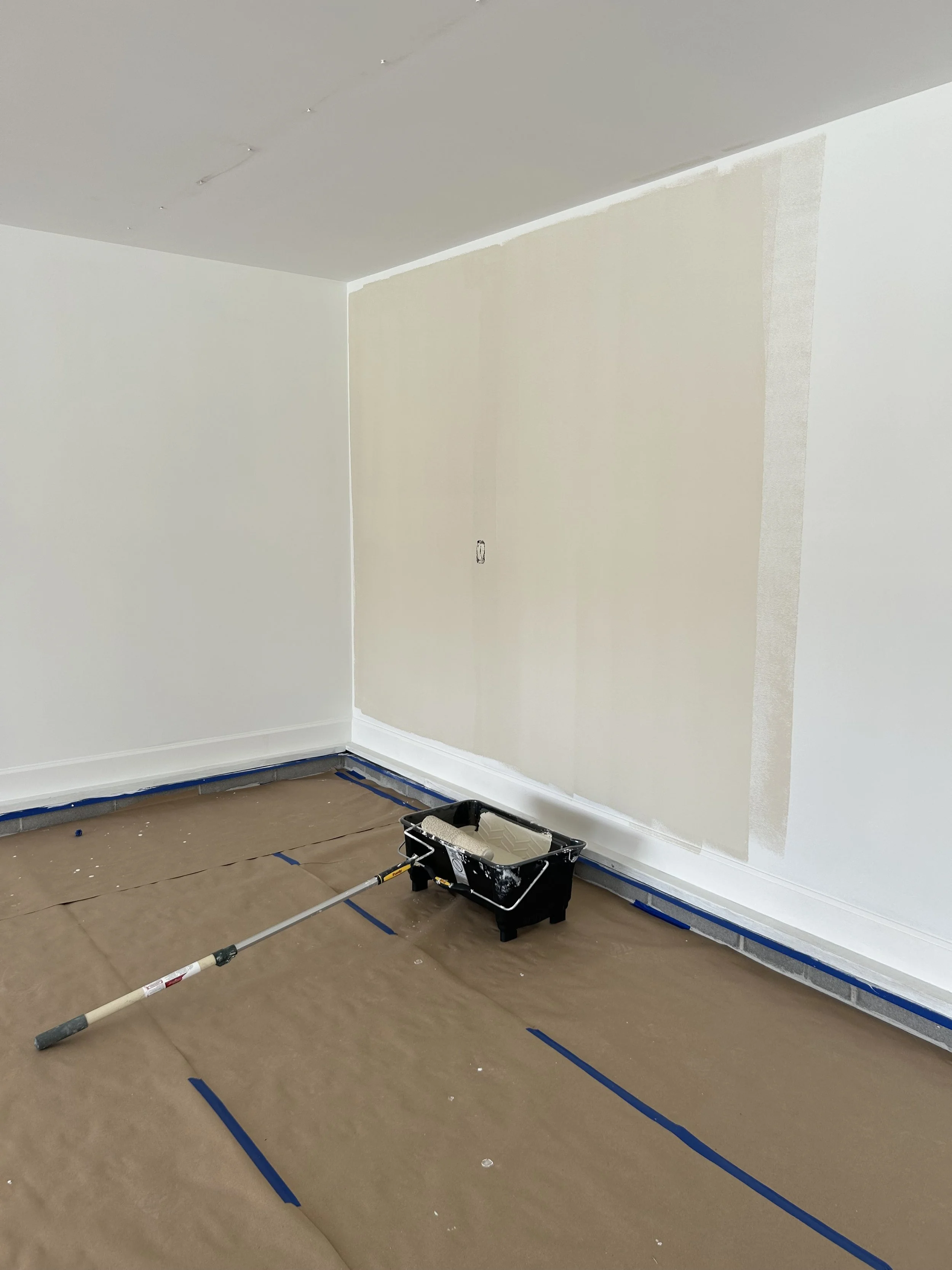 Room under renovation with a beige wall partially painted, a container of white paint, and brown paper covering the floor.
