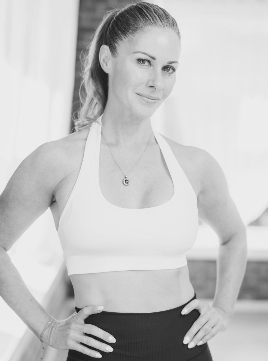 A woman with a high ponytail wearing a white sports bra and black leggings, standing with hands on hips, smiling at the camera in a bright room.