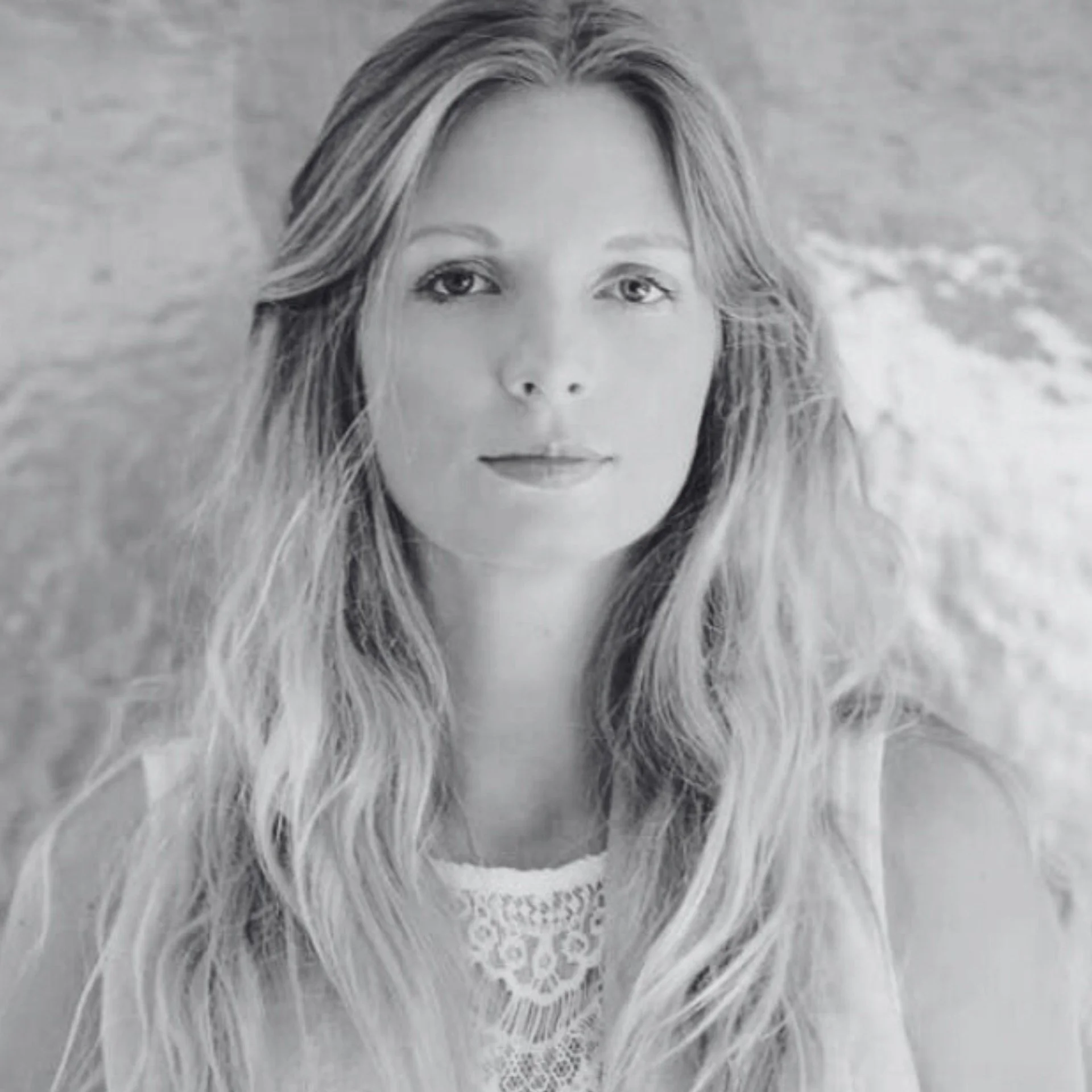 Black-and-white photo of a woman's face with long hair, looking directly at the camera, with some lace fabric underneath.