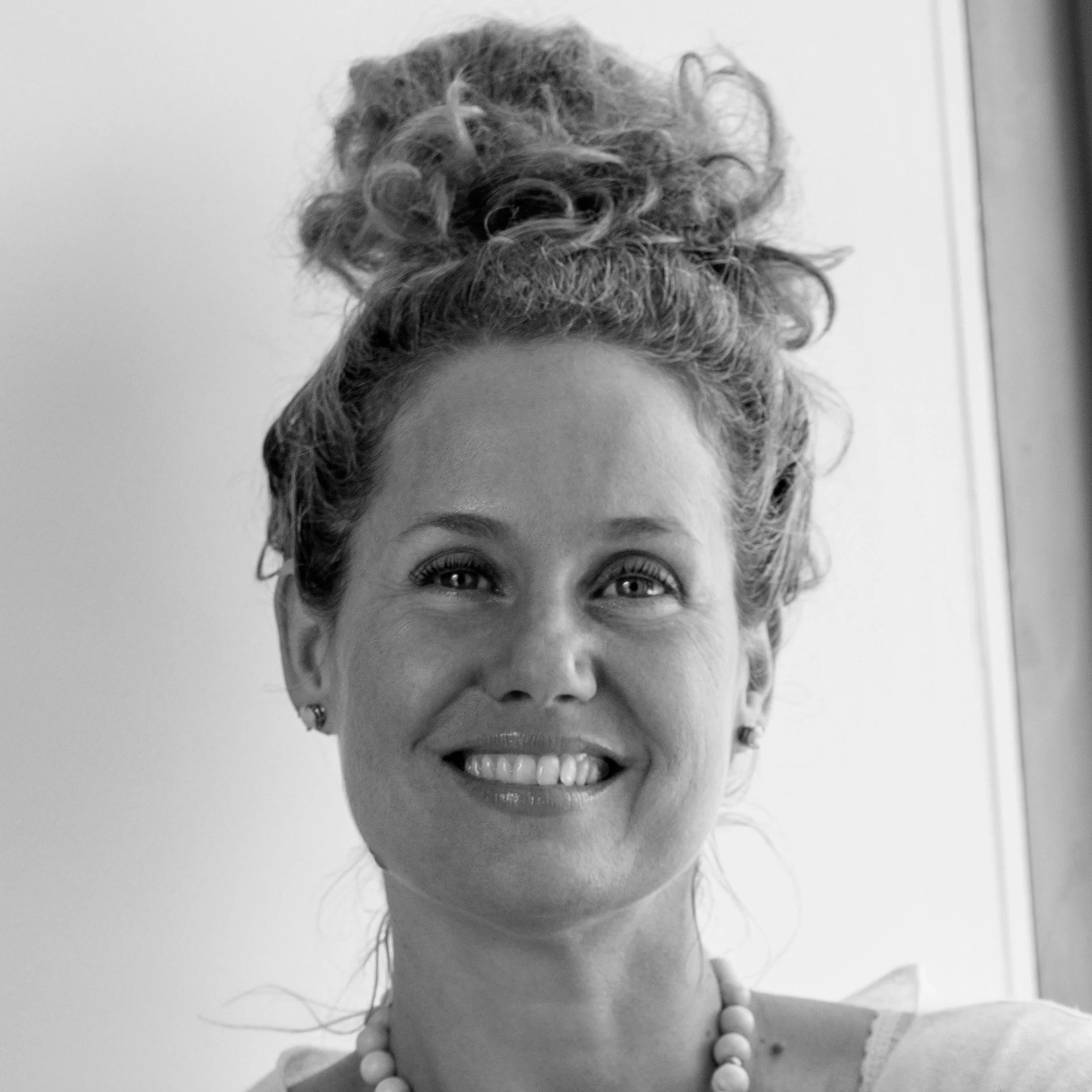 Black and white photo of smiling woman with curly hair styled in an updo, wearing pearl necklace and earrings.