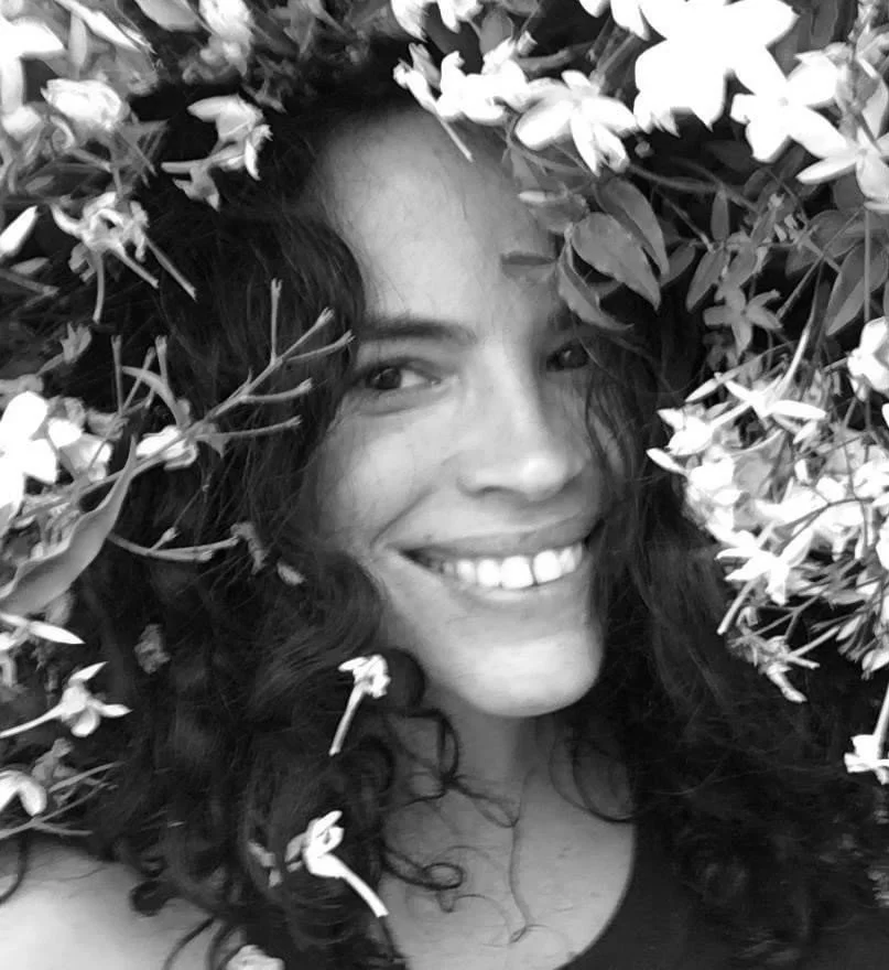 A woman smiling with dark, curly hair surrounded by flowers.