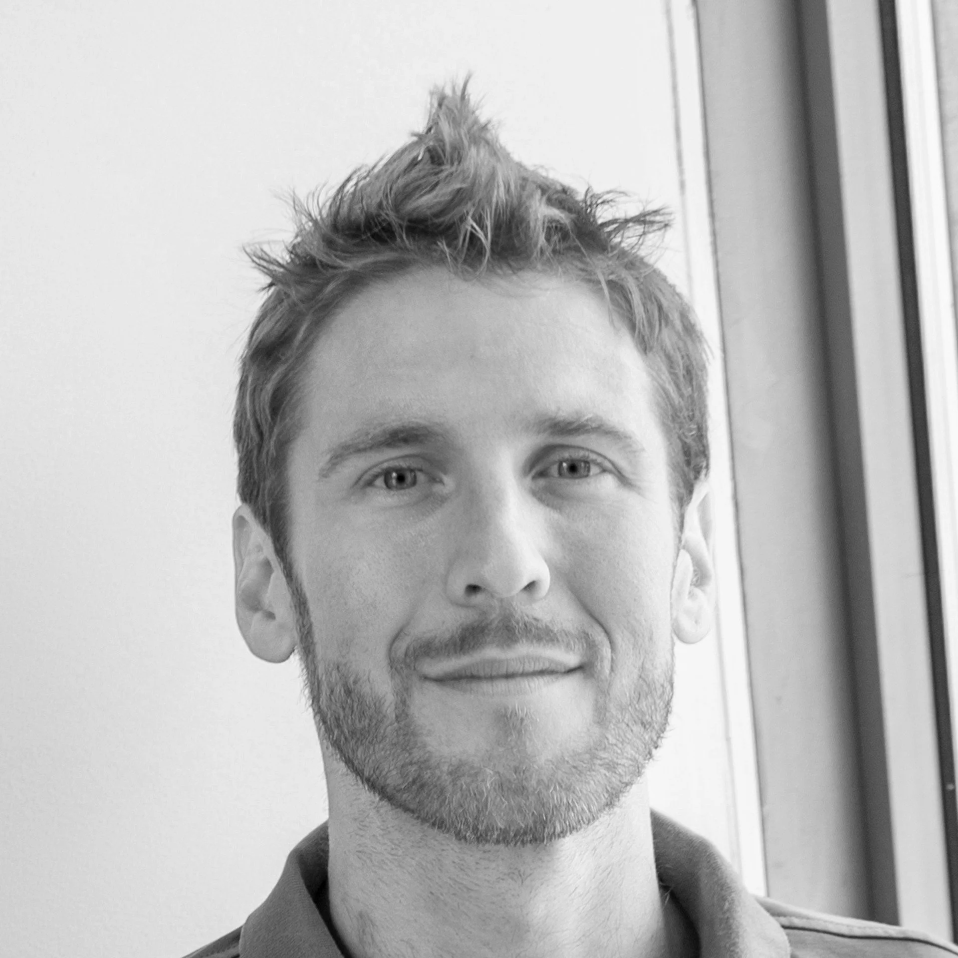 Close-up black and white photo of a person with short, spiky hair and visible beard and mustache, looking directly at the camera, standing indoors near a door or window.