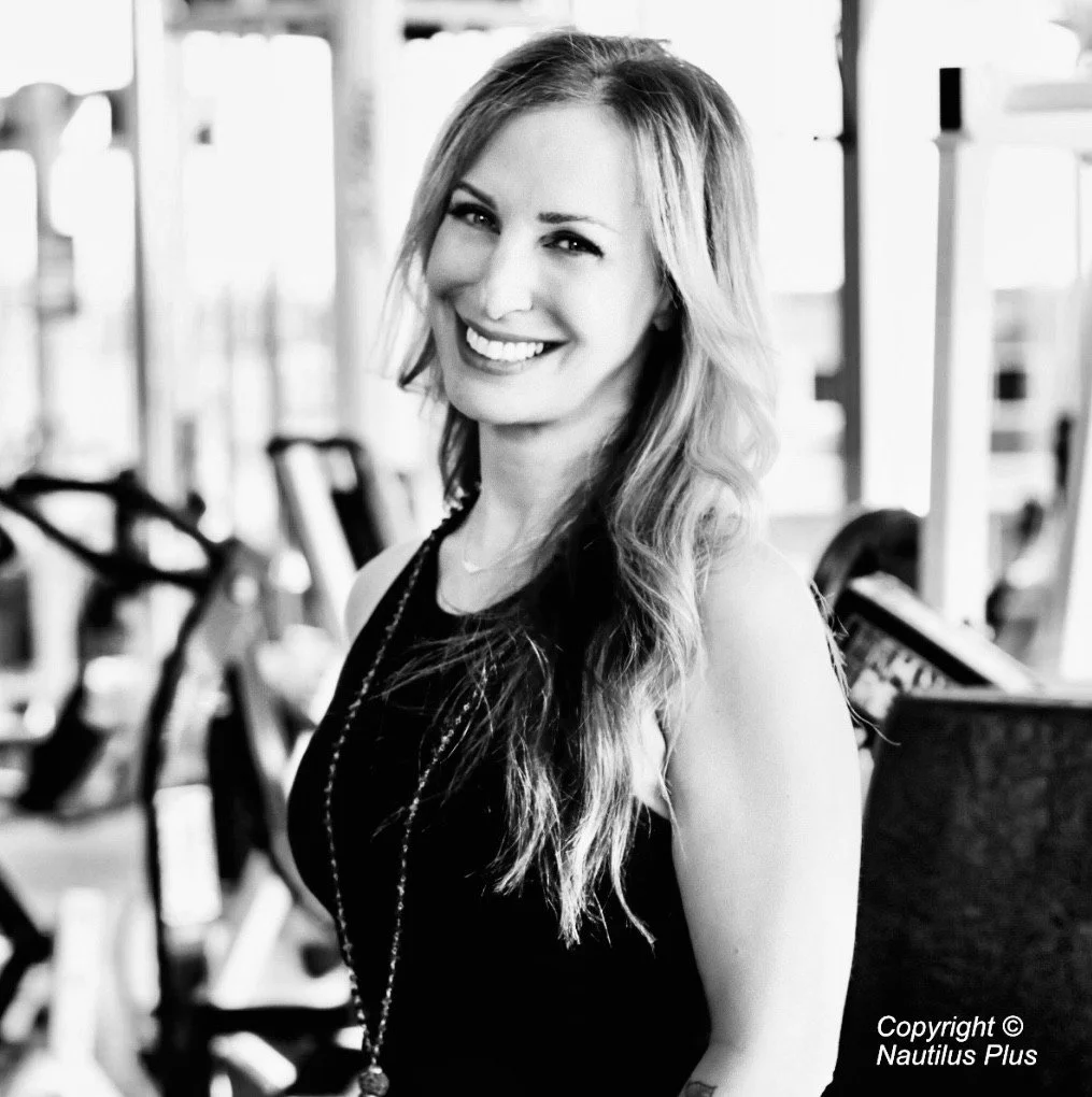 A smiling woman with long wavy hair, wearing a sleeveless top, standing in a gym with exercise equipment in the background.