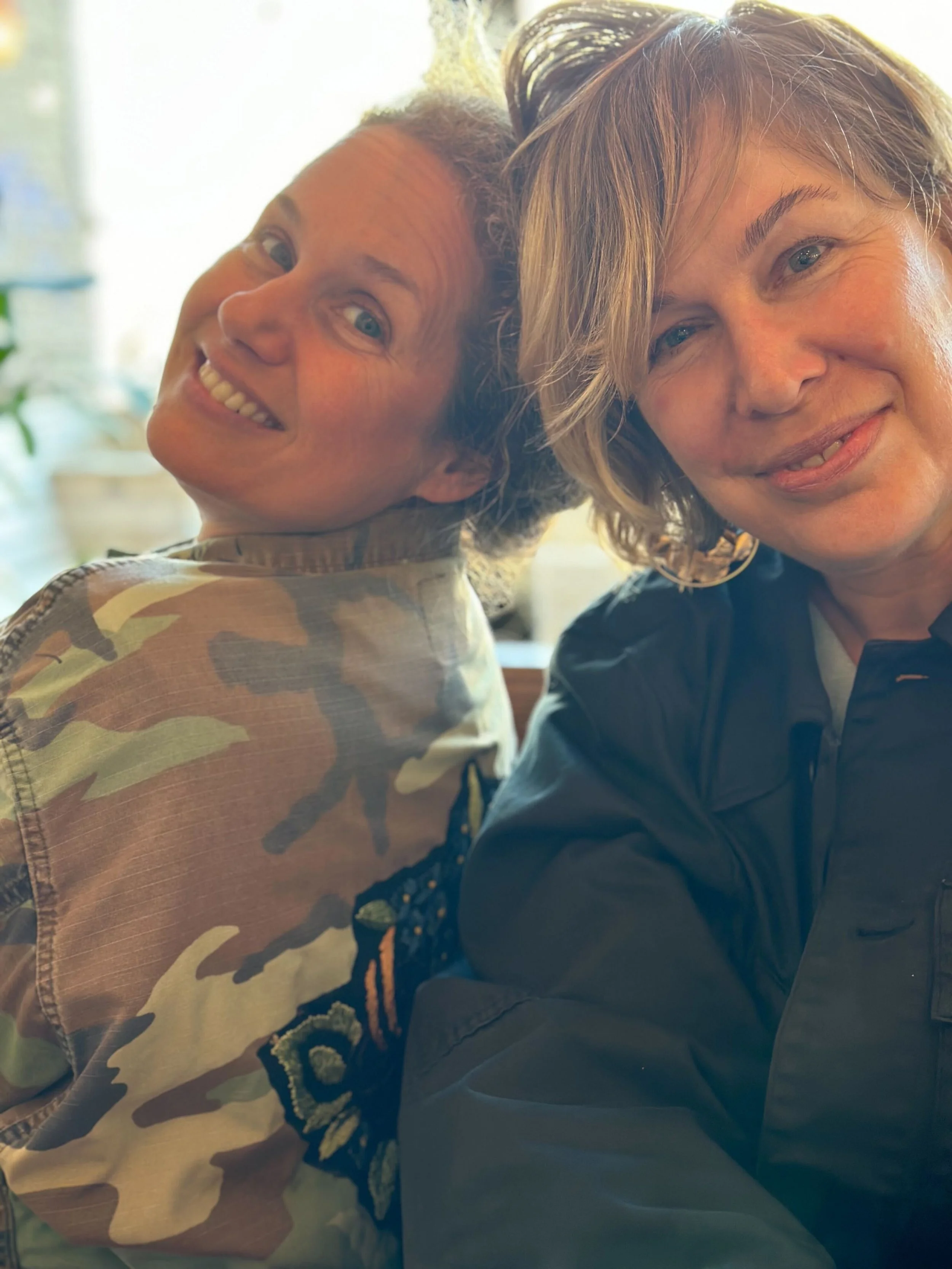 A woman with short, wavy blonde hair and a woman with curly hair smiling and leaning their heads together in an outdoor setting.