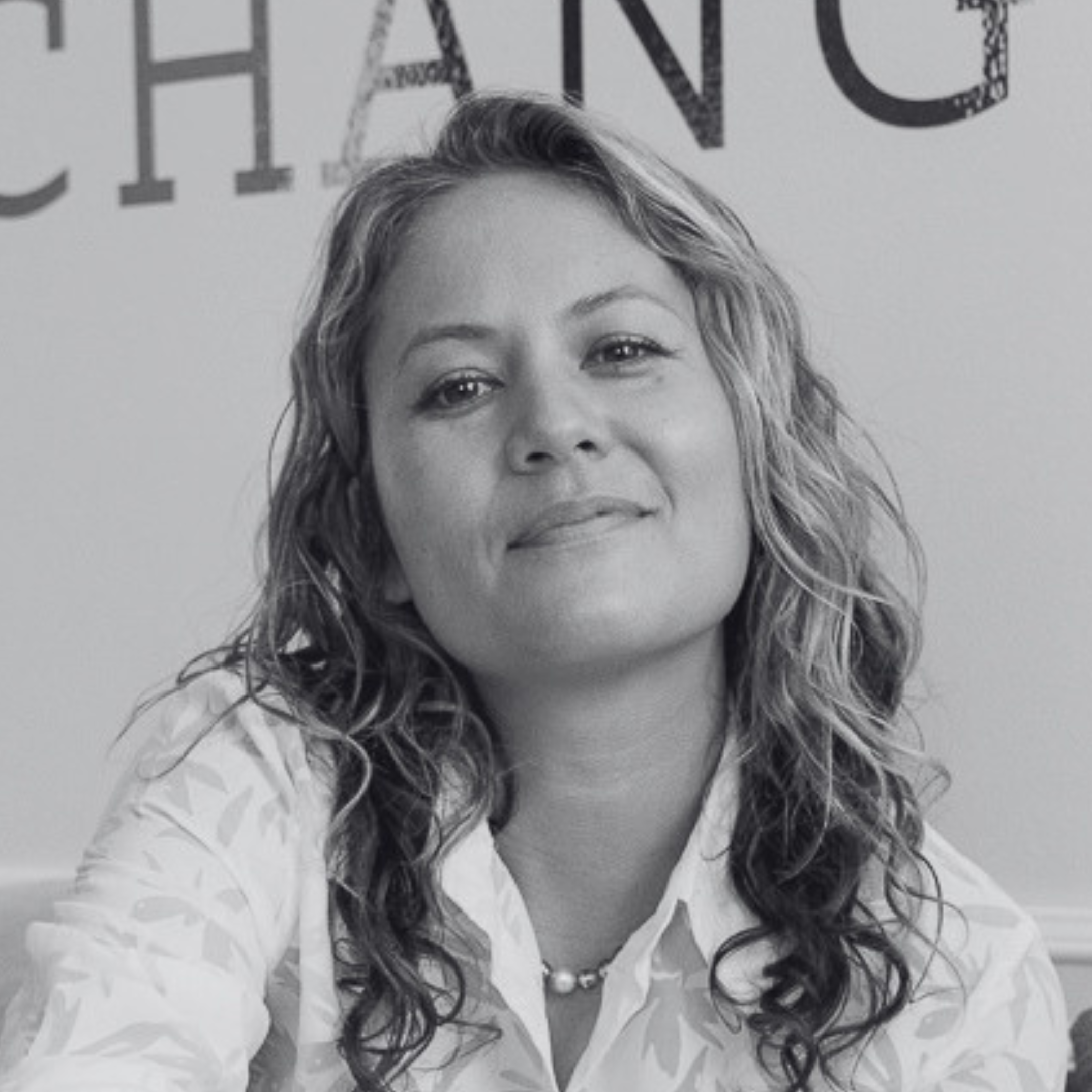 Black and white portrait of a woman with wavy hair, wearing a necklace with pearls, standing in front of a wall with partially visible text.