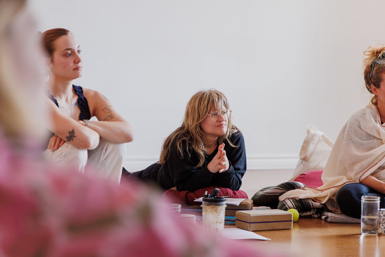 Women sitting on the floor with notebooks, drinks, and personal items, attentively listening during a meeting or workshop in a bright room.