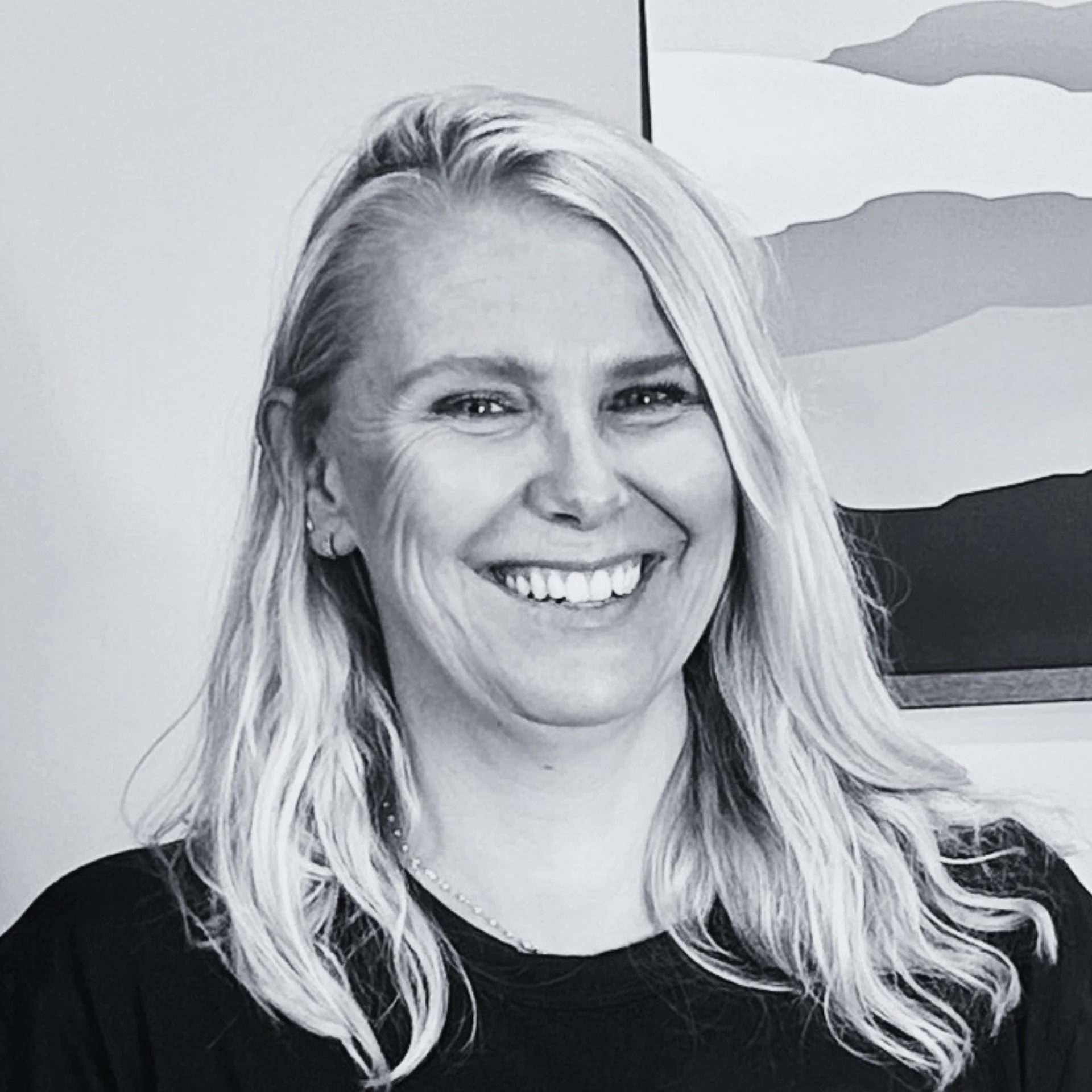 Black and white portrait photograph of a smiling woman with long, wavy hair, wearing jewelry, and a dark top, with a mountain-like abstract design in the background.