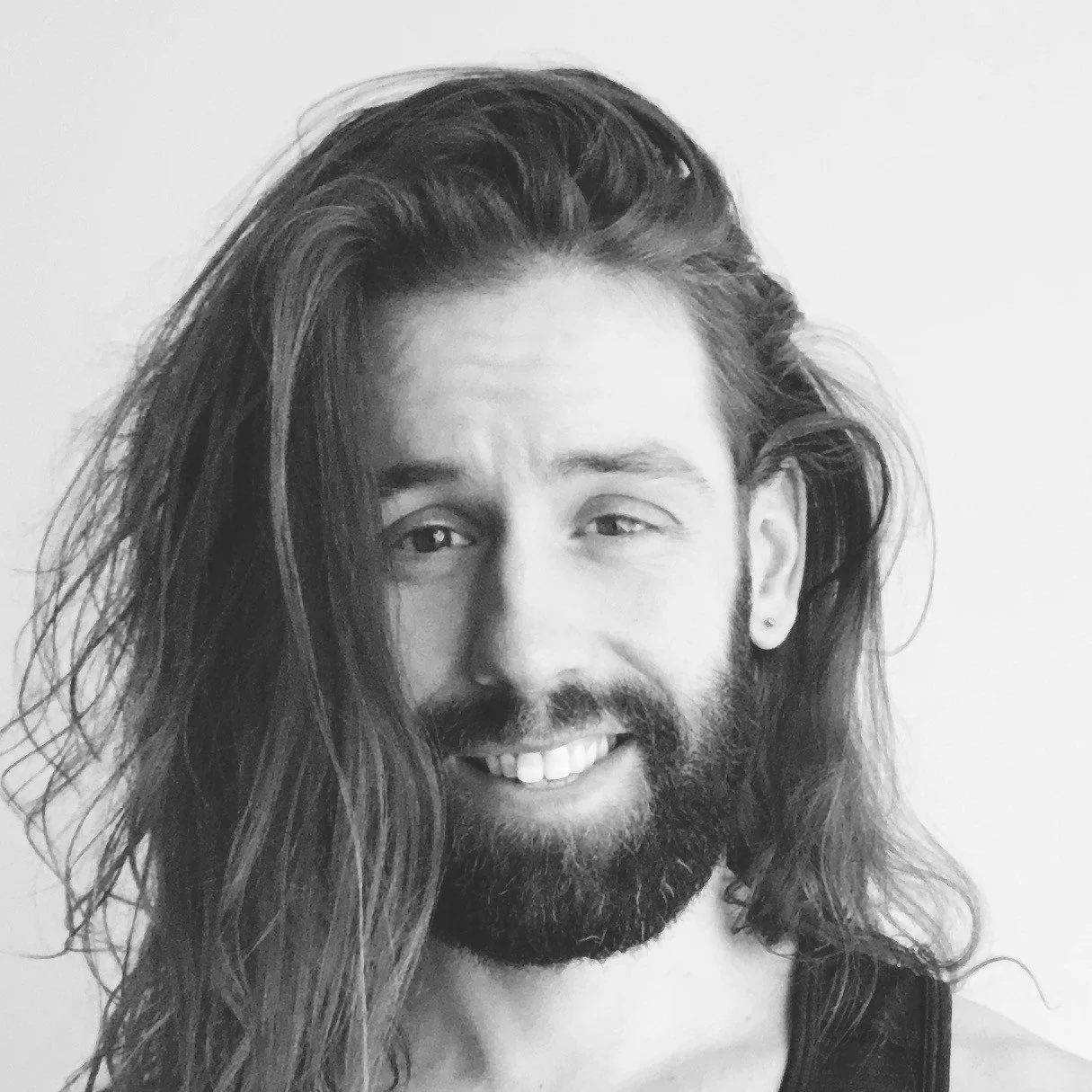 Black and white photo of a young man with long wavy hair, a beard, and a small earring in his left ear, smiling gently.