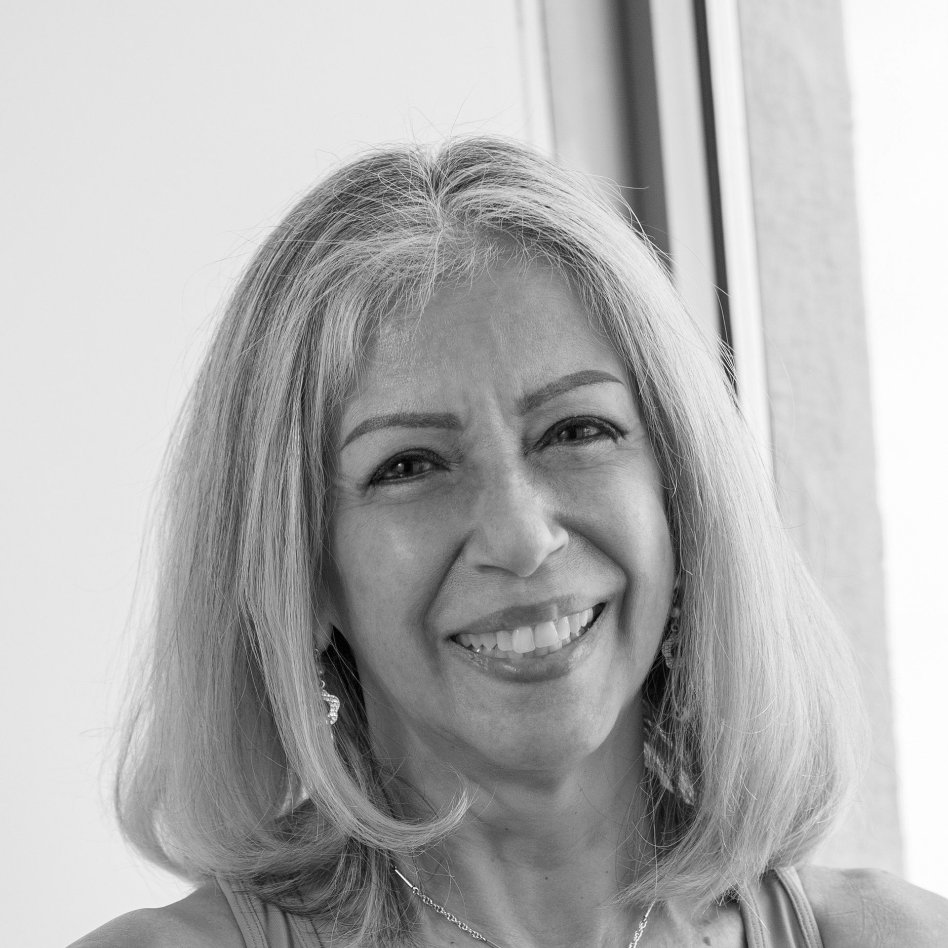 Black and white photo of a smiling woman with blonde hair, wearing earrings and a necklace, near a window.
