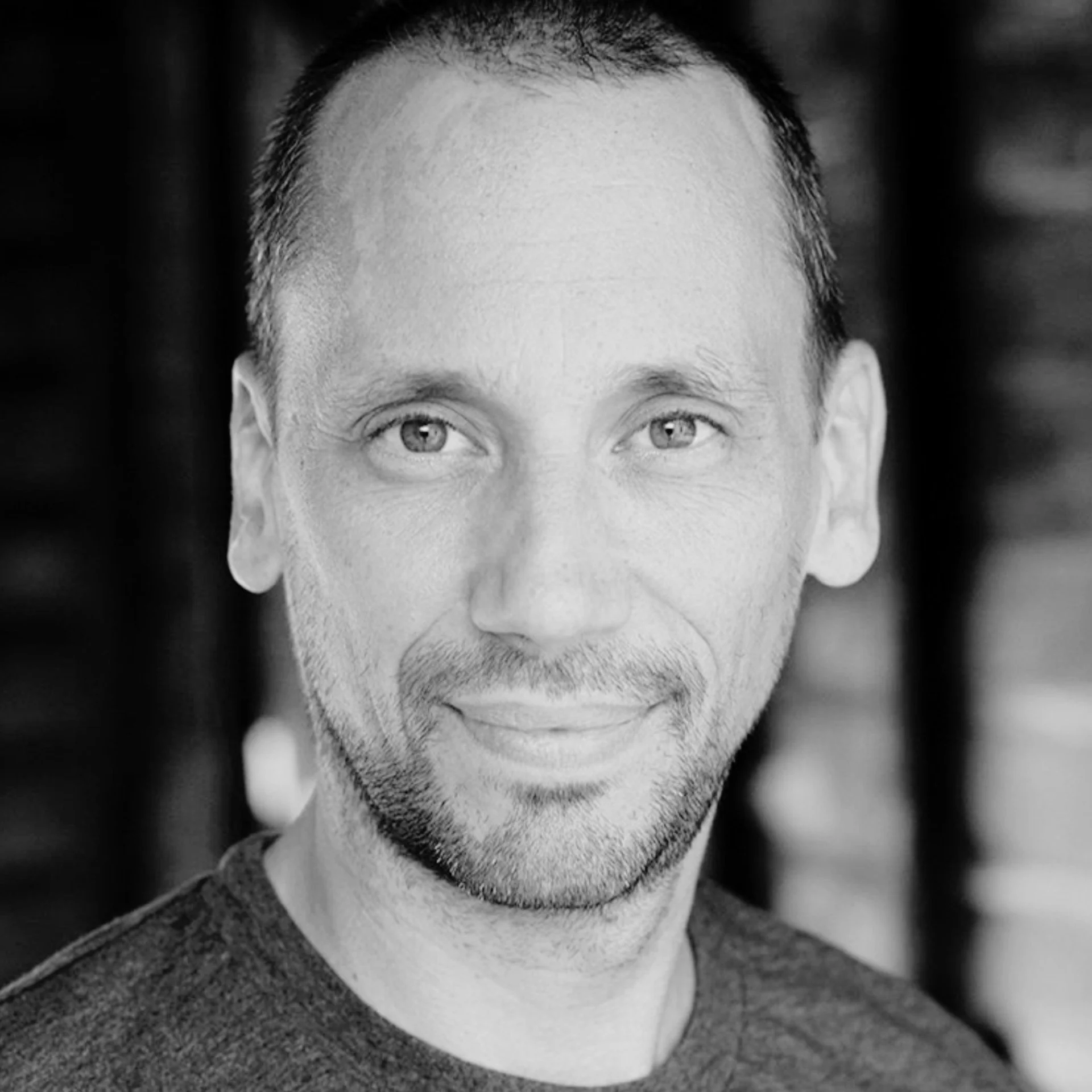 Black and white photo of a man's face, partially visible, showing eyes, nose, and beard, with short hair, wearing a hoodie.