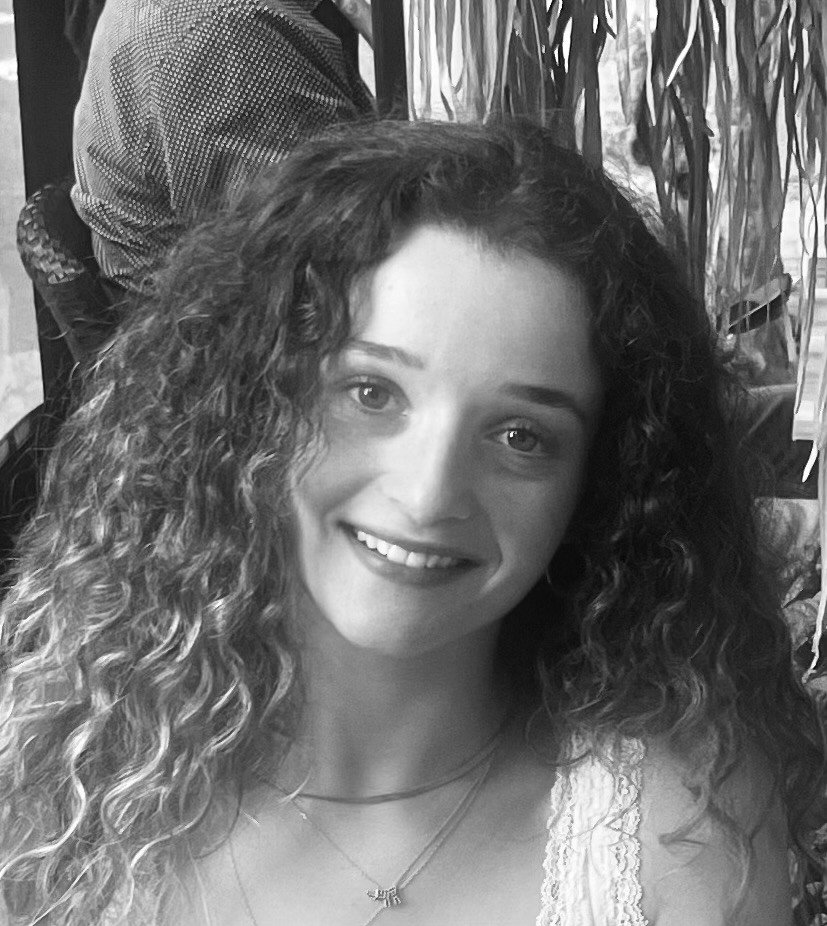 A young woman with long curly hair smiling at the camera, wearing a necklace, and sitting in a room with indoor plants.