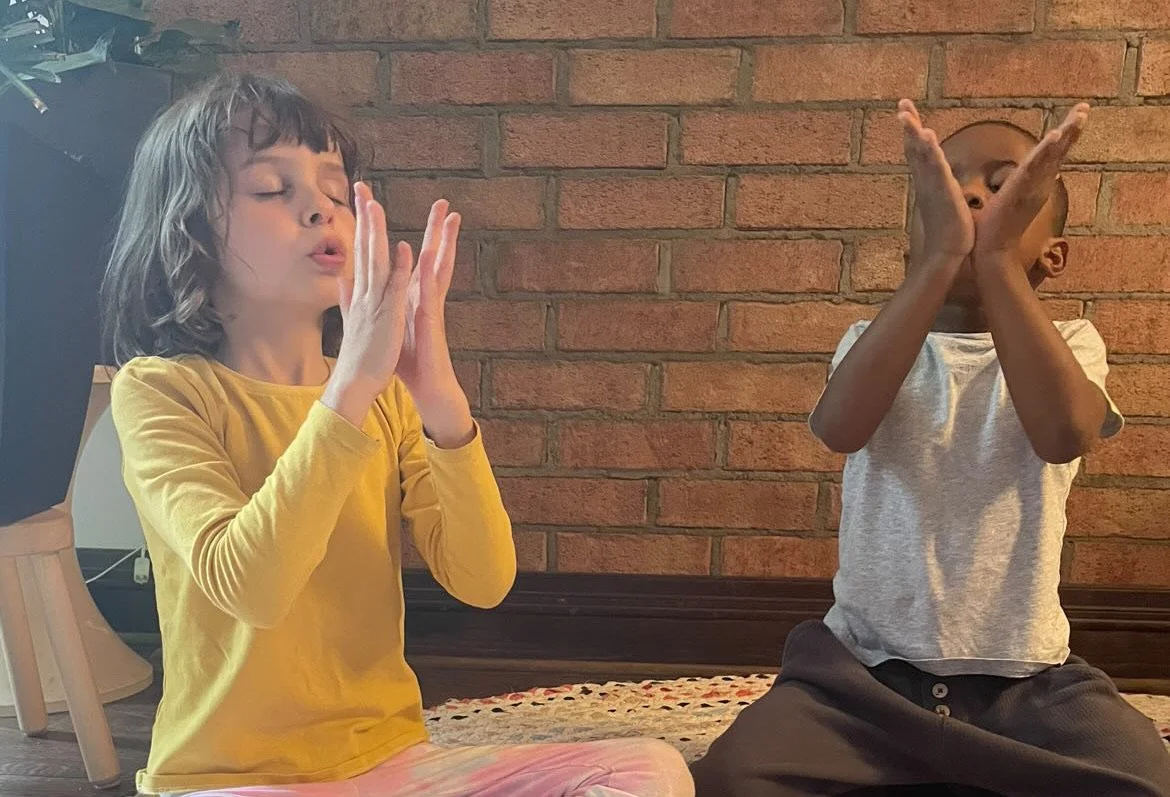 Two children sitting on the floor with their eyes closed, making blowing or breathing gestures with their hands crossed in front of their faces, in front of a brick wall.