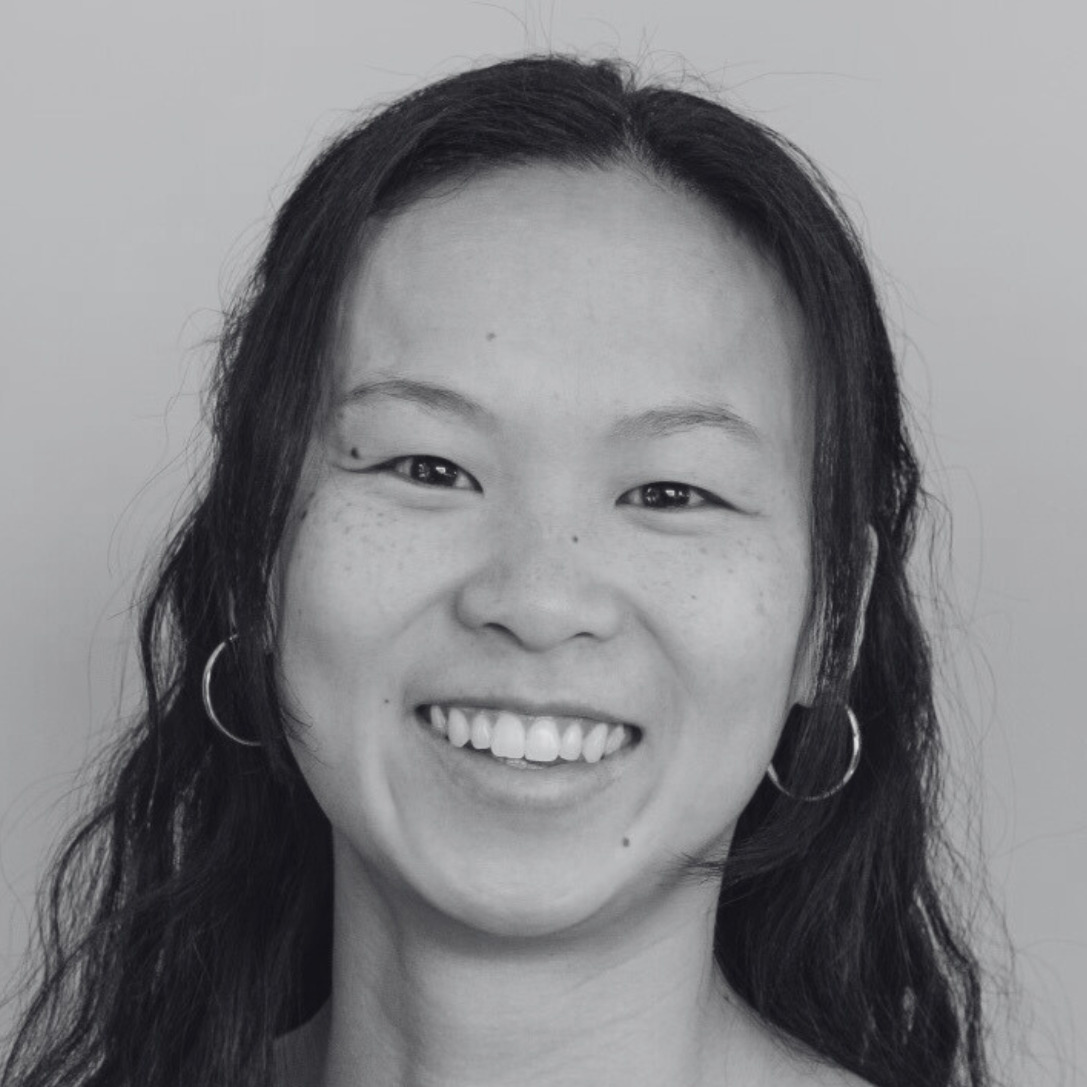 Black and white close-up of a smiling woman with dark hair, wearing hoop earrings, and a light-colored background.