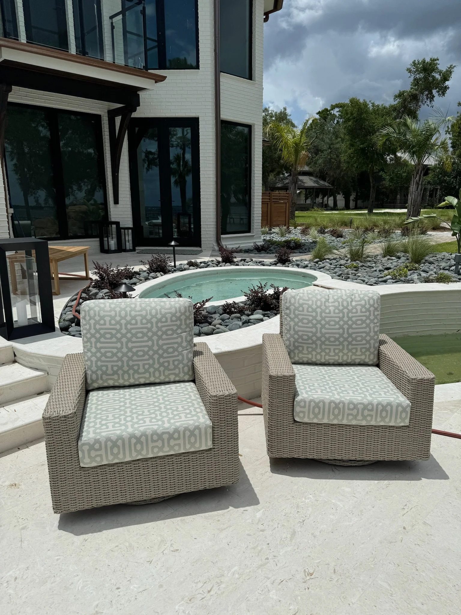 Outdoor patio area with two wicker chairs with patterned cushions, a small round hot tub, paver stones, and landscaped garden with trees and plants near modern white brick house with large glass windows and an enclosed balcony.