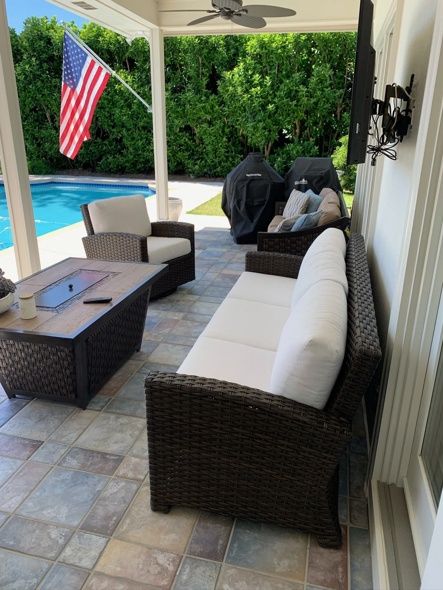Patio with wicker furniture, a beige sofa, cushioned chairs, a table with a candle and remote, a ceiling fan, a flag hanging, a swimming pool beyond a grassy area, green bushes in the background, and an electric grill covered with a black cover.