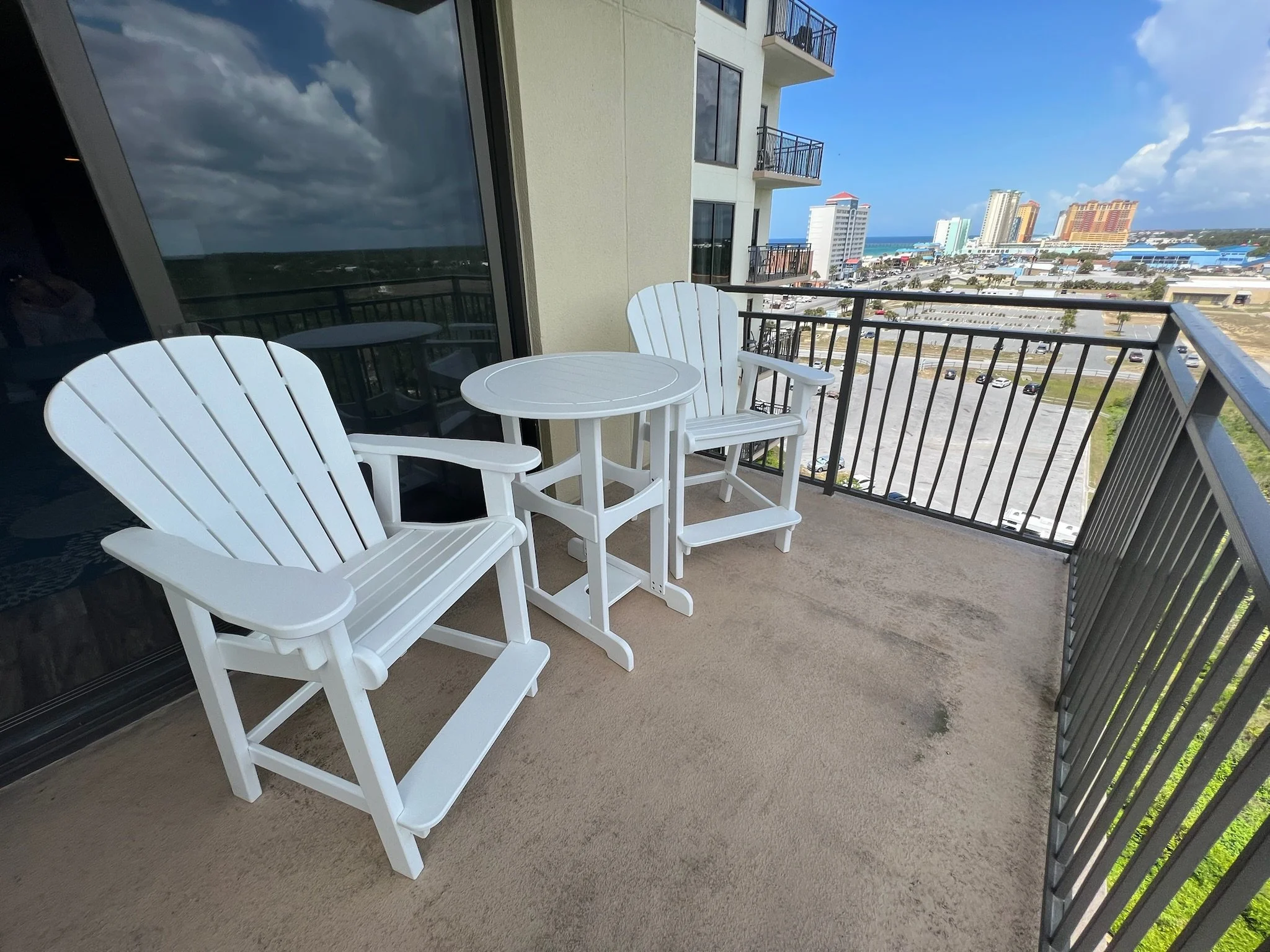 Balcony with white chairs and table overlooking a cityscape with high-rise buildings and a beach in the background.