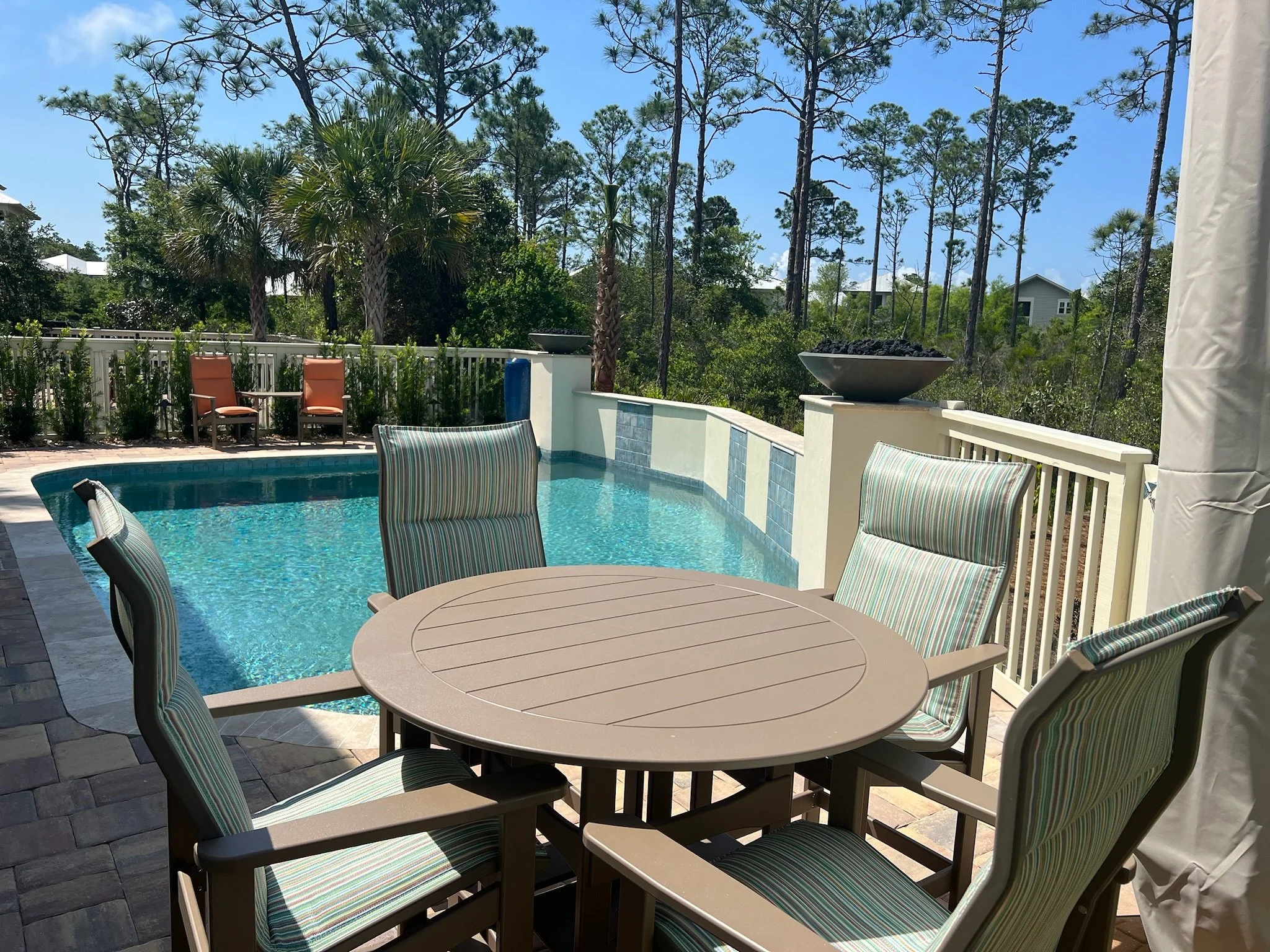 A backyard patio with a round table and chairs near a swimming pool, surrounded by a white fence and lush green trees with a blue sky in the background.