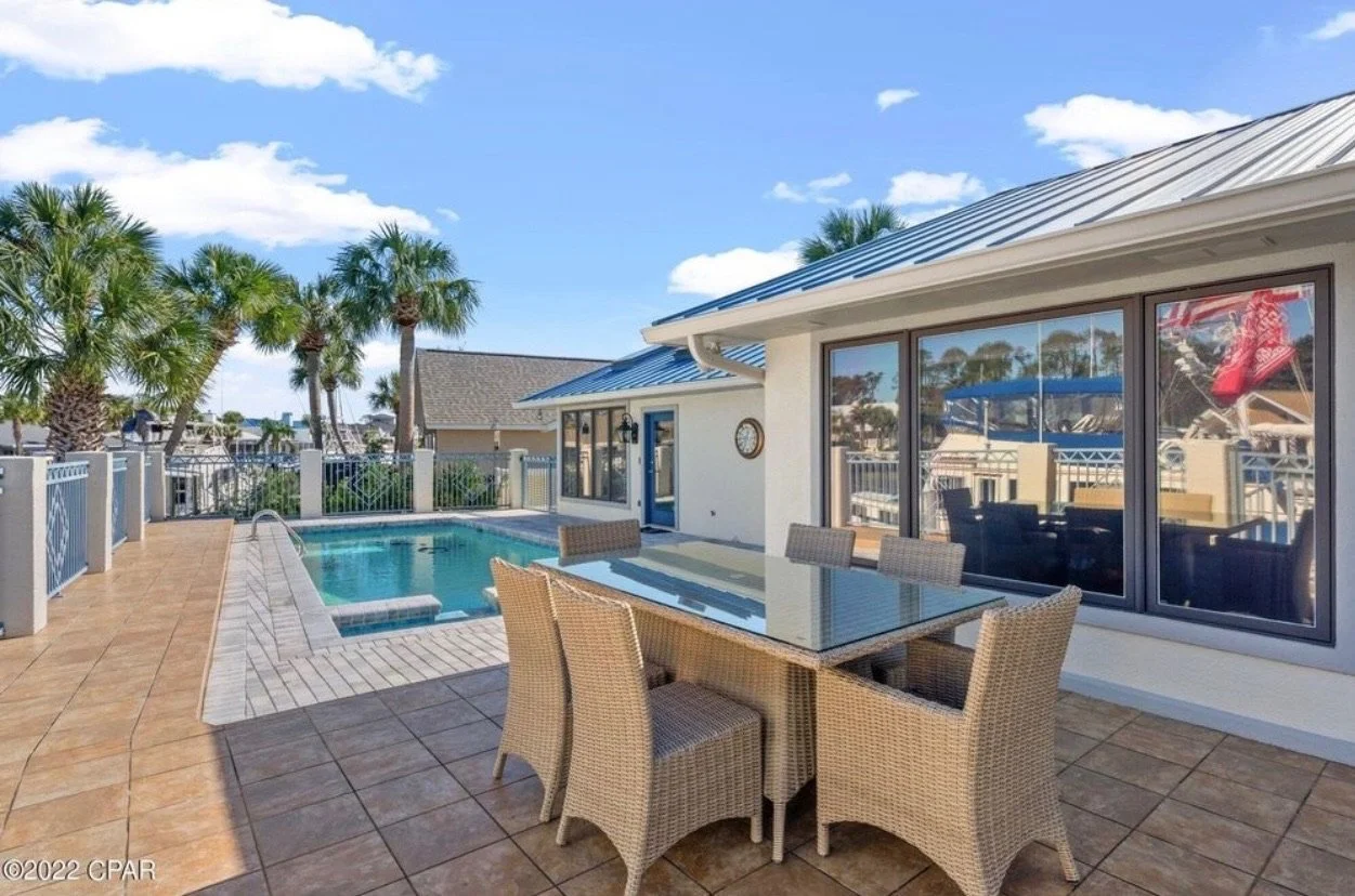 Residential backyard patio with a swimming pool, outdoor dining table with six chairs, and palm trees, under a partly cloudy blue sky.